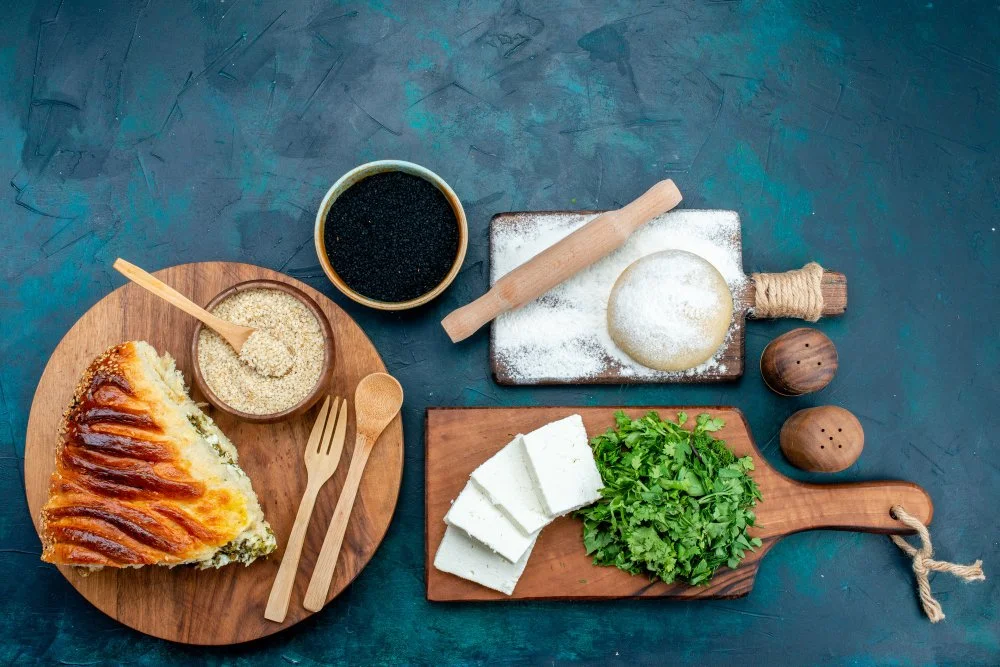 A wooden plate with a slice of spinach and cheese pie, a small bowl of sesame seeds, and wooden fork and spoon. Nearby, a bowl of black sesame seeds, a rolling pin, a round piece of dough, and two wooden peppercorn shakers. Also, a wooden cutting board with chopped white cheese and fresh cilantro.