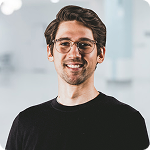 A man with curly brown hair, glasses, and a beard, smiling at the camera, wearing a black shirt in an indoor setting.