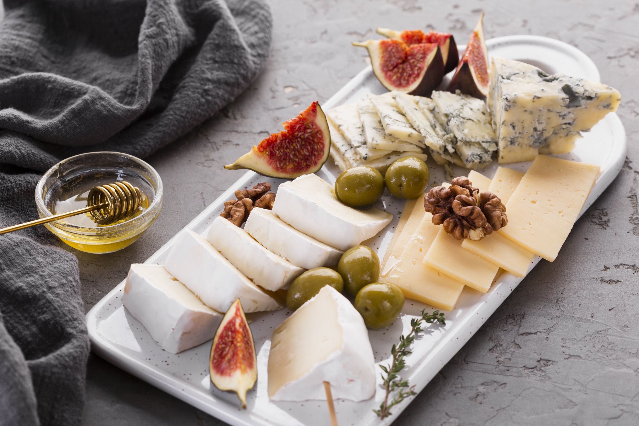 A cheese platter featuring a variety of cheeses, green olives, fresh figs, walnuts, and blue cheese on a white rectangular plate. Honey in a small glass bowl with a honey dipper is beside the plate, and a gray cloth is draped nearby.
