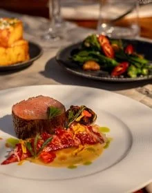A plate with a cooked steak topped with herbs, served with a vegetable medley and a colorful sauce. In the background, there is a bowl of mashed potatoes and a black bowl of mixed greens salad on a dining table.