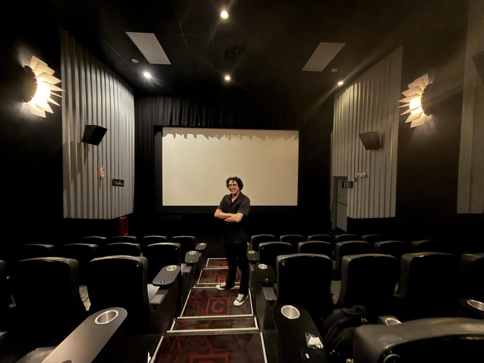 Una sala de cine oscura con una pantalla y dos personas, una en el centro y otra en la esquina, que parecen estar preparándose para la película.