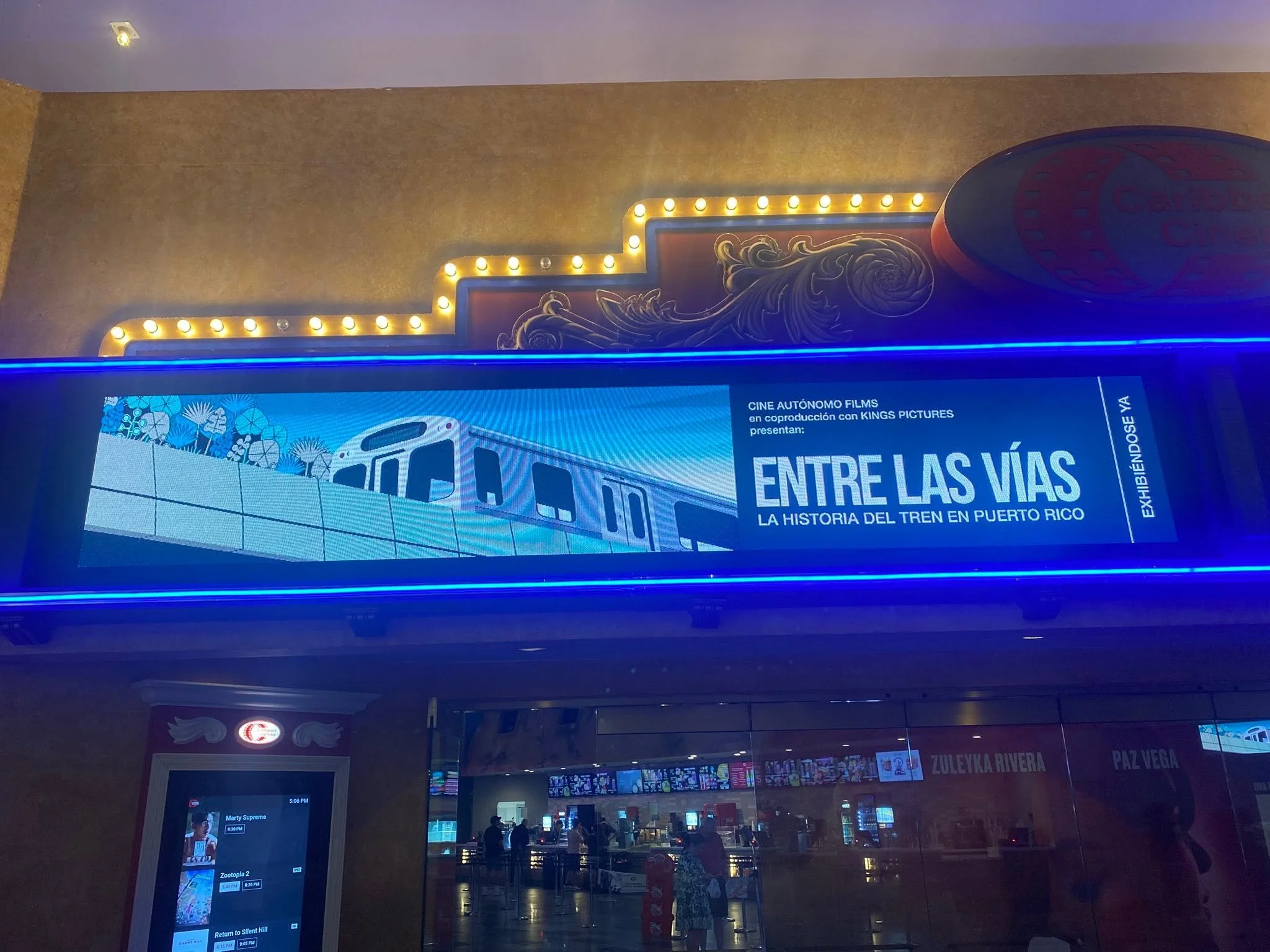 Pantalla digital en un cine con información sobre películas, en un entorno oscuro con luces decorativas amarillas y azules, y personas en el fondo.