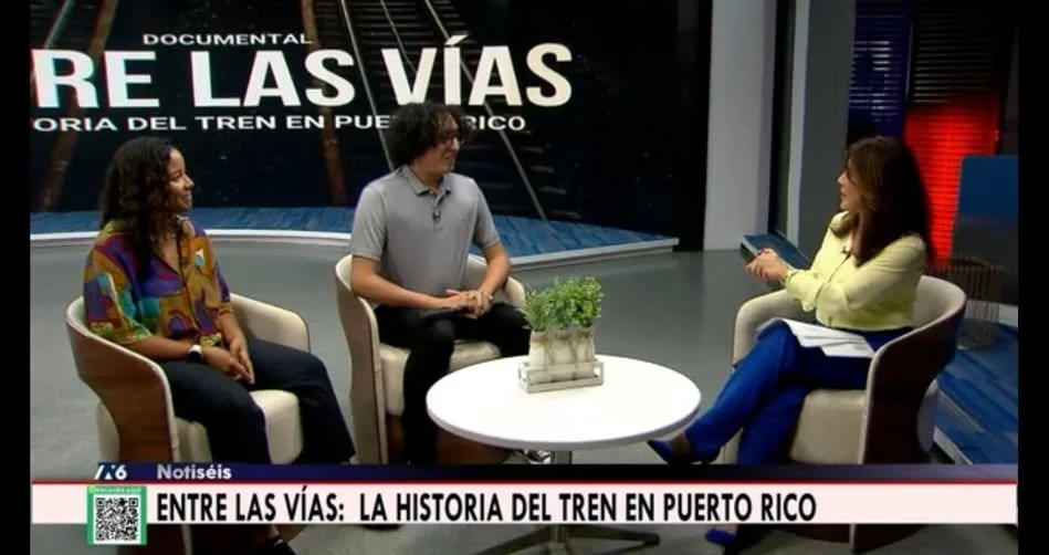 Tres personas en un programa de televisión discutiendo sobre la historia del tren en Puerto Rico, con un cartel que dice 'RE LAS VÍAS' y un subtítulo que dice 'Entre las vías: La historia del tren en Puerto Rico'.