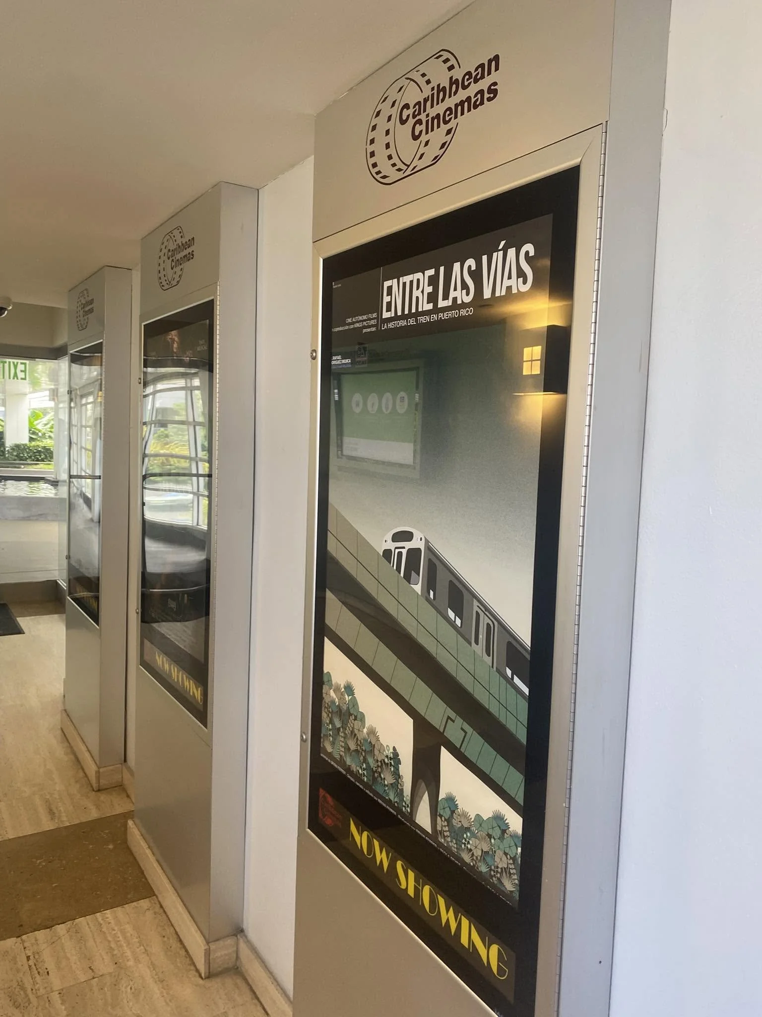 Carteles y pantalla informativa en un pasillo con señales de cine Caribbean Cinemas y un cartel que dice "Entre las vías" y "Now Showing"