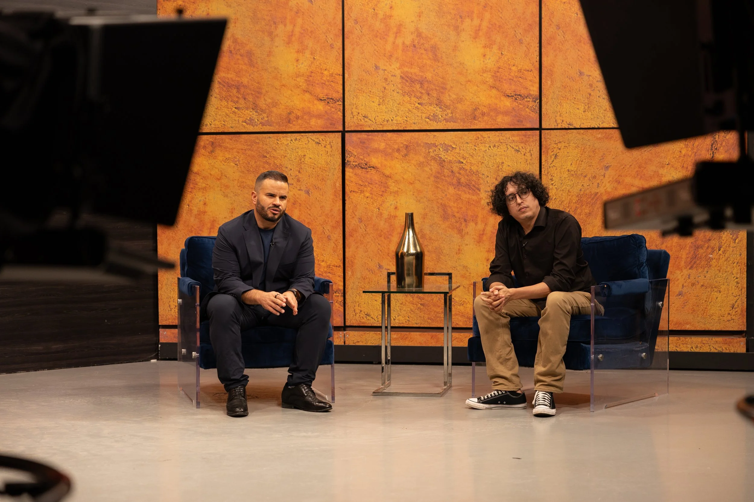 Dos hombres sentados en una entrevista o conversación en un escenario con fondo anaranjado, con cámaras alrededor observando.