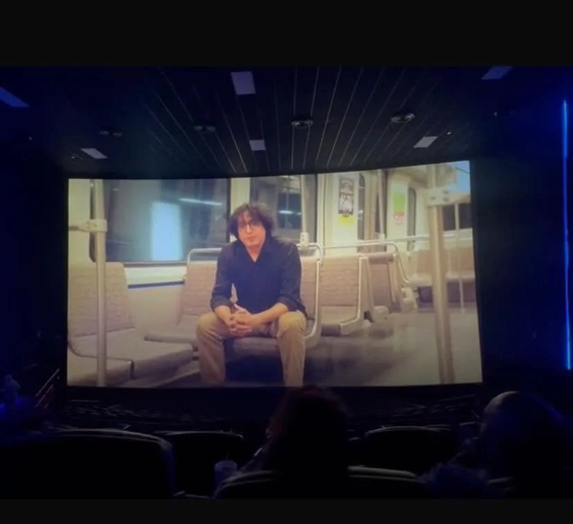 Una pantalla grande en un cine mostrando a una persona sentada en un vagón del metro.