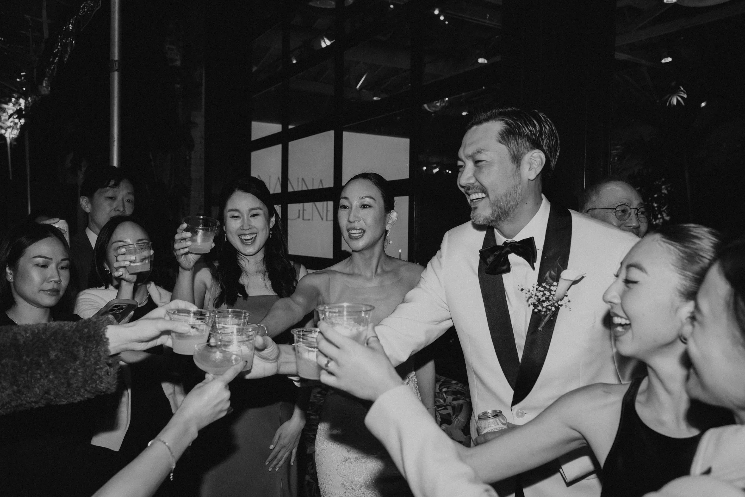 Black and white photo of bride and groom cheering their drinks with a bunch of their guest