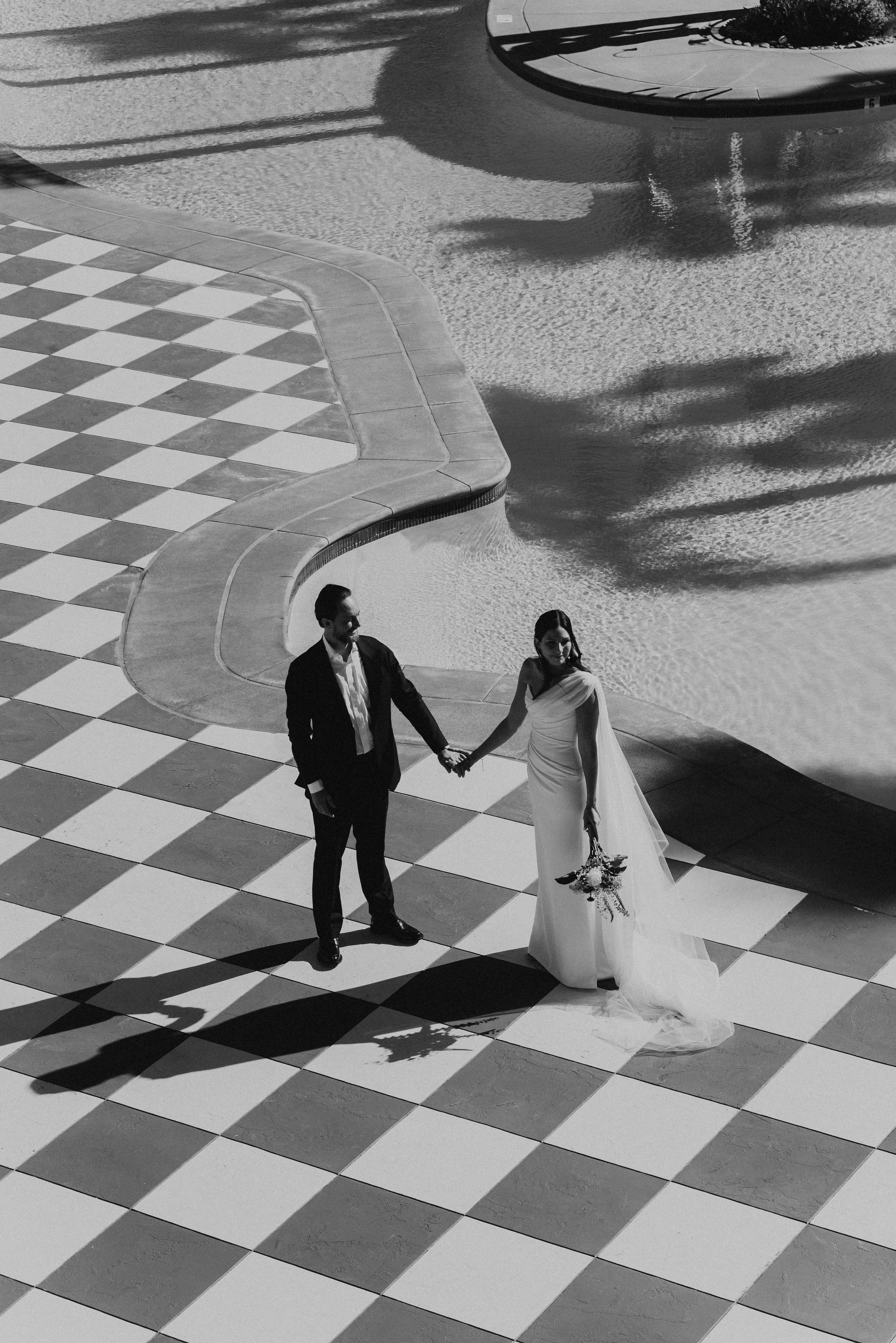 black and white photo of Bride and groom standing holding hands on checkered floor with pool behind them