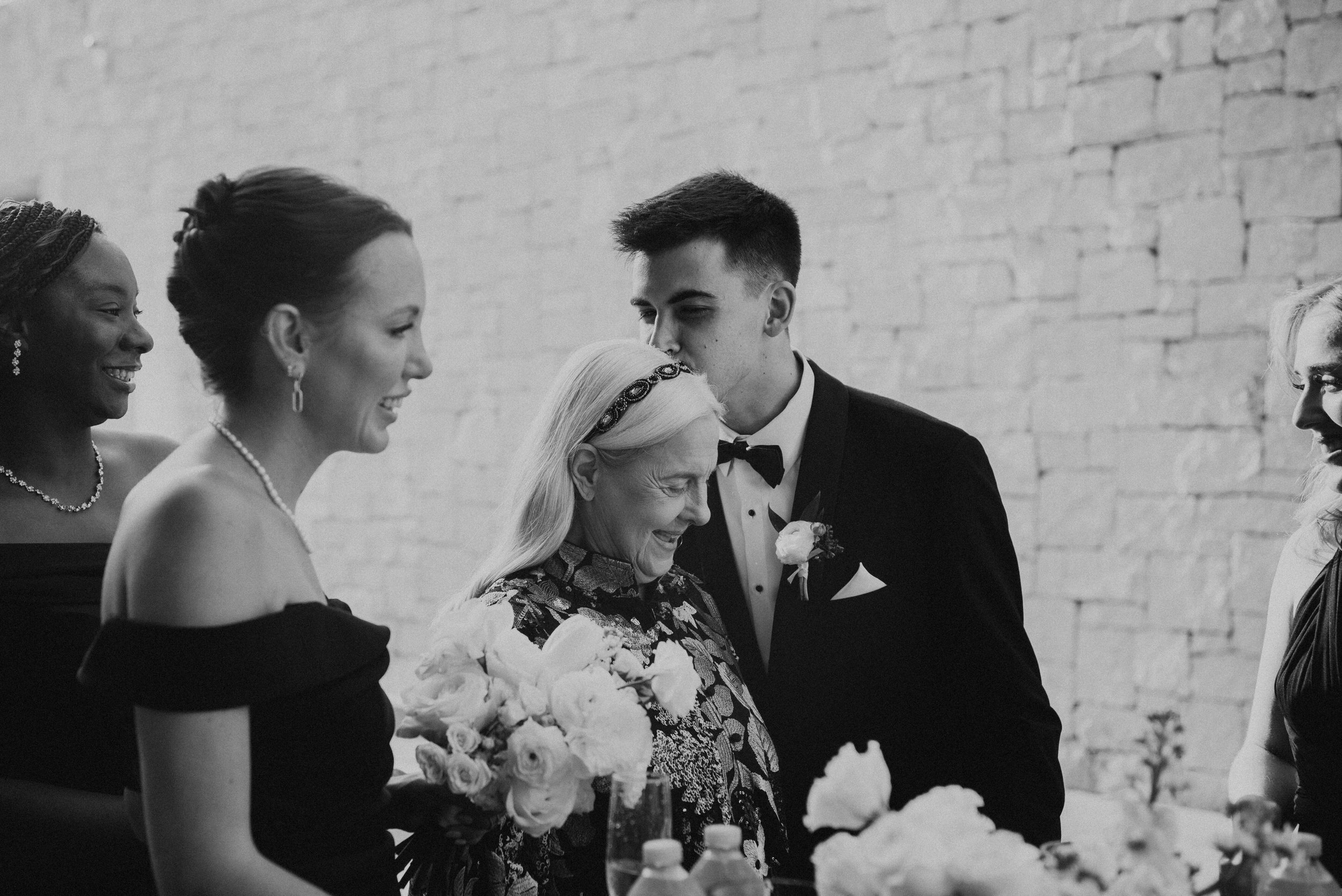 black and white photo of man in suit kissing his grandmother on the side of her head with other smiling in the photo around them