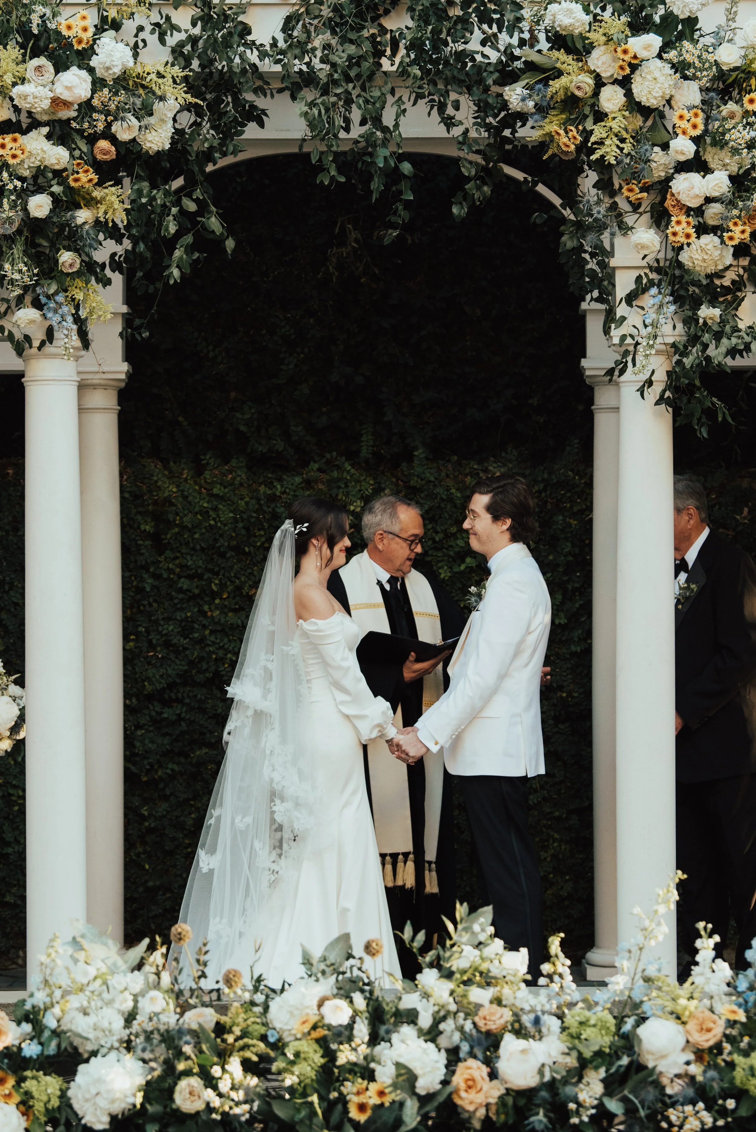 close up of Bride and groom facing each other holding hand at their ceremony. You can see the flowers covering the white arbor at the top of the photo.