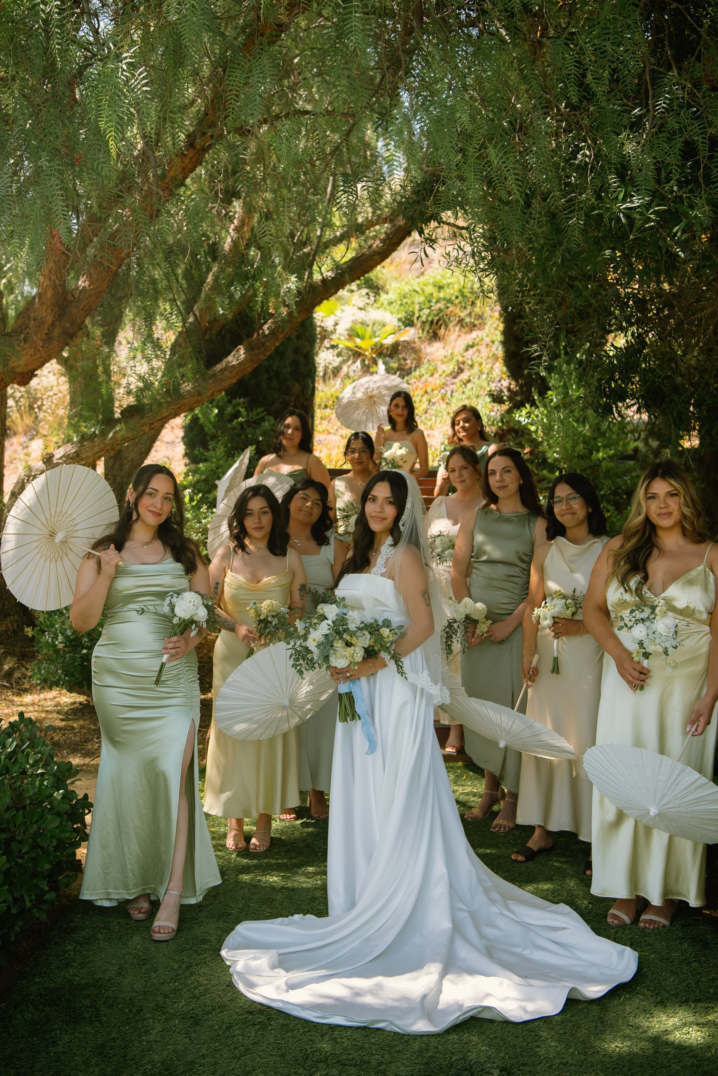 bride and bridesmaid posing for the camera under some tress. Some of the bridesmaids are holding white umbrellas.
