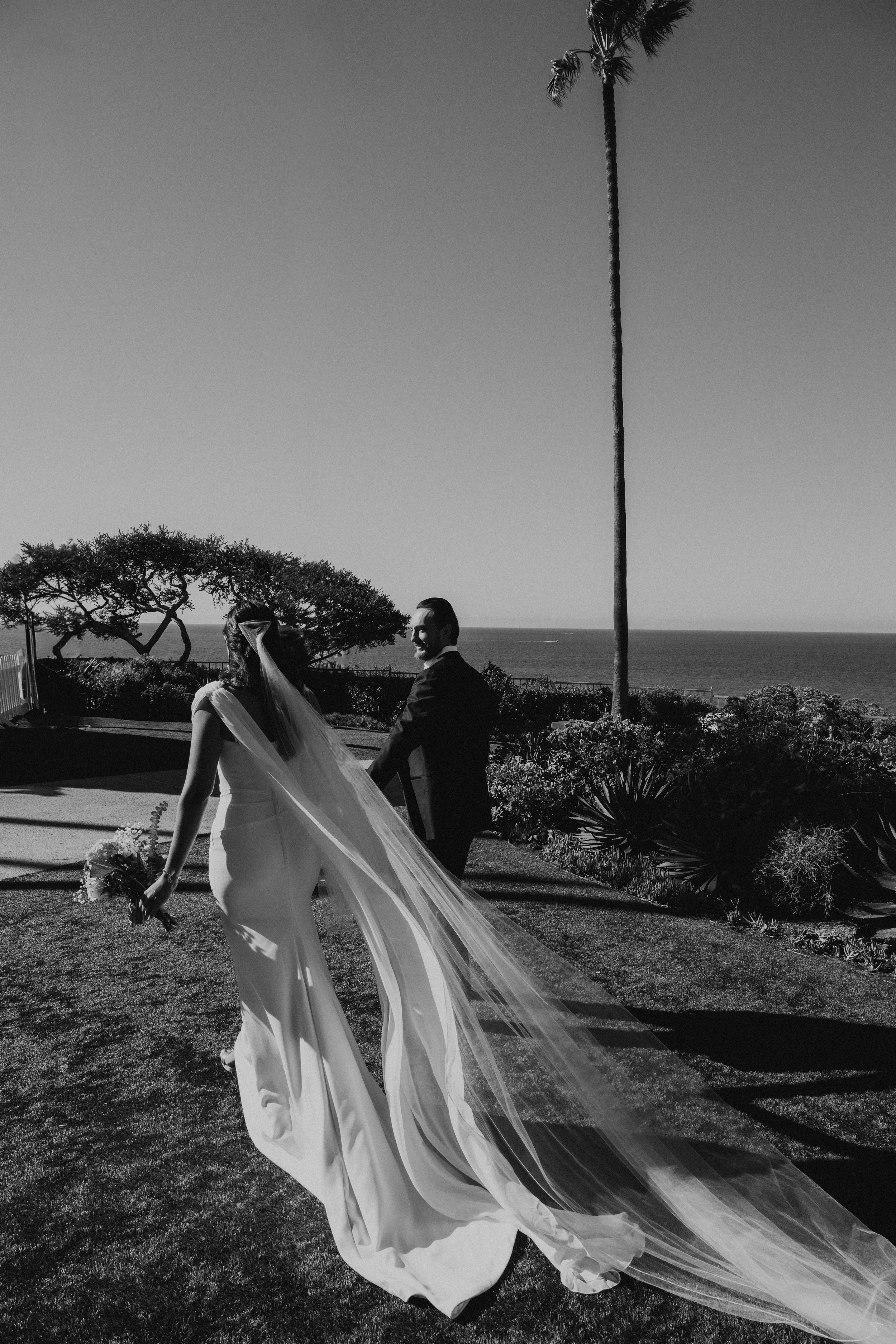 Black and white photo of bride and groom walking away from the camera. Brides dress and veil is trailing behind her