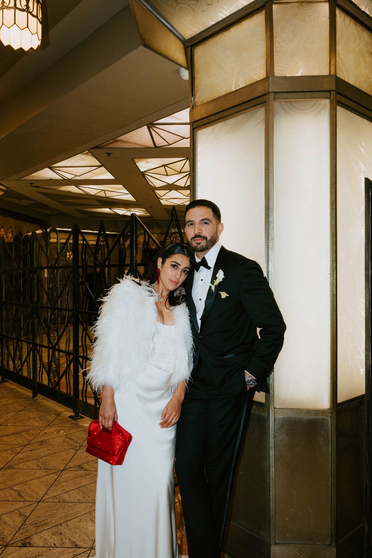 Groom leaning against a lit up wall looking at the camera and bride is leaning on his chest also looking at camera. Both are looking serious at the camera.