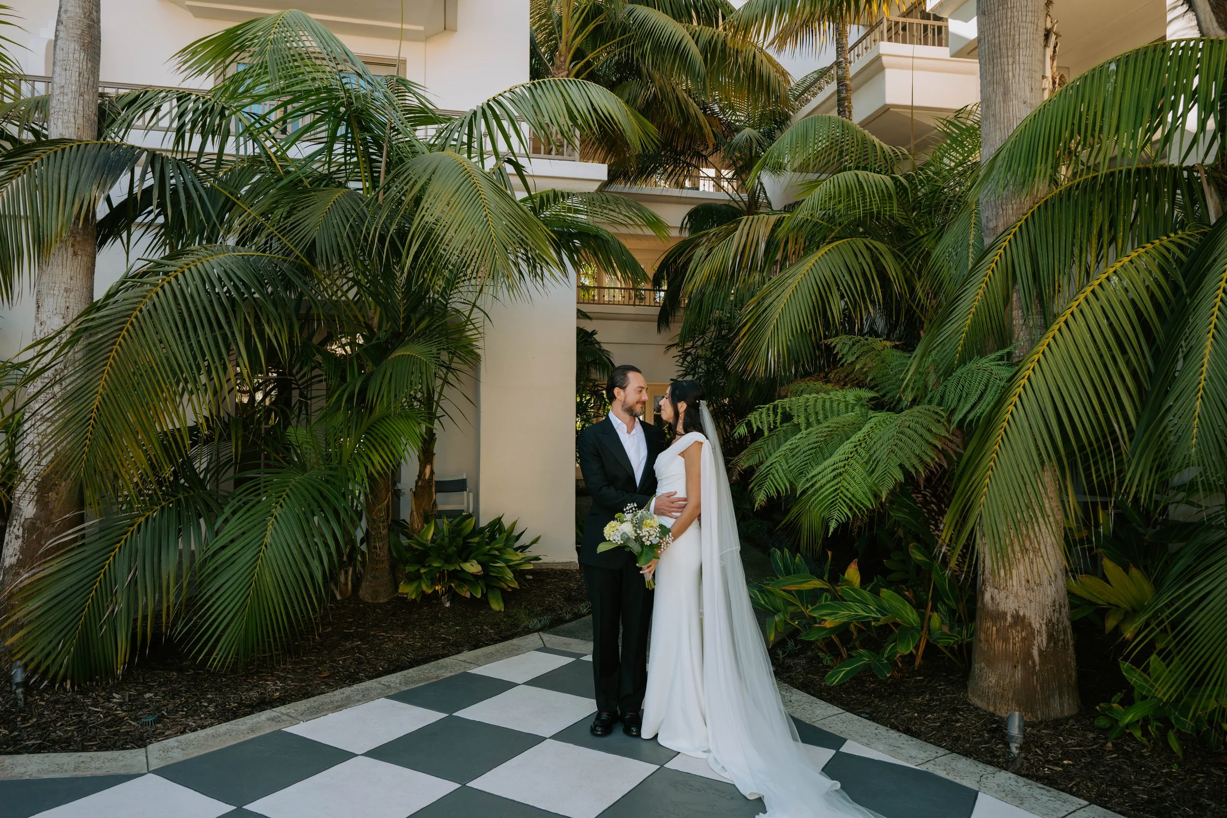 Bride and groom standing on a checkered floor surrounded by trees looking at each other