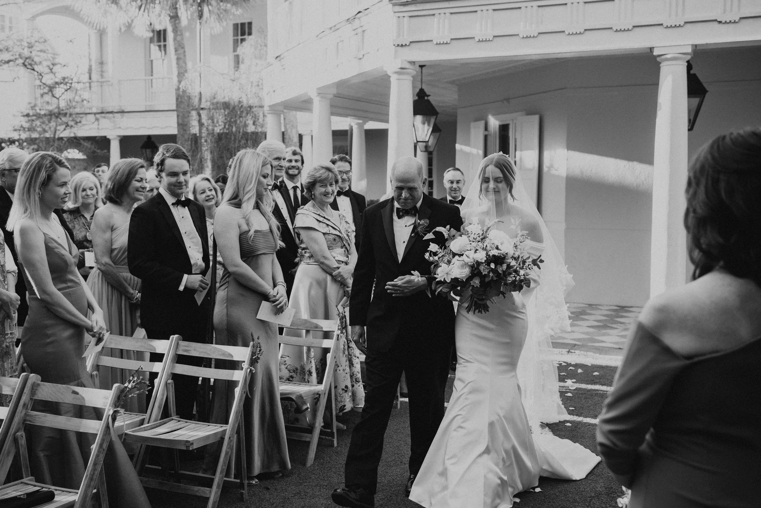 Black and white photo of Bride and her father walking up he aisle. You can see guest smiling while standing at their seats.