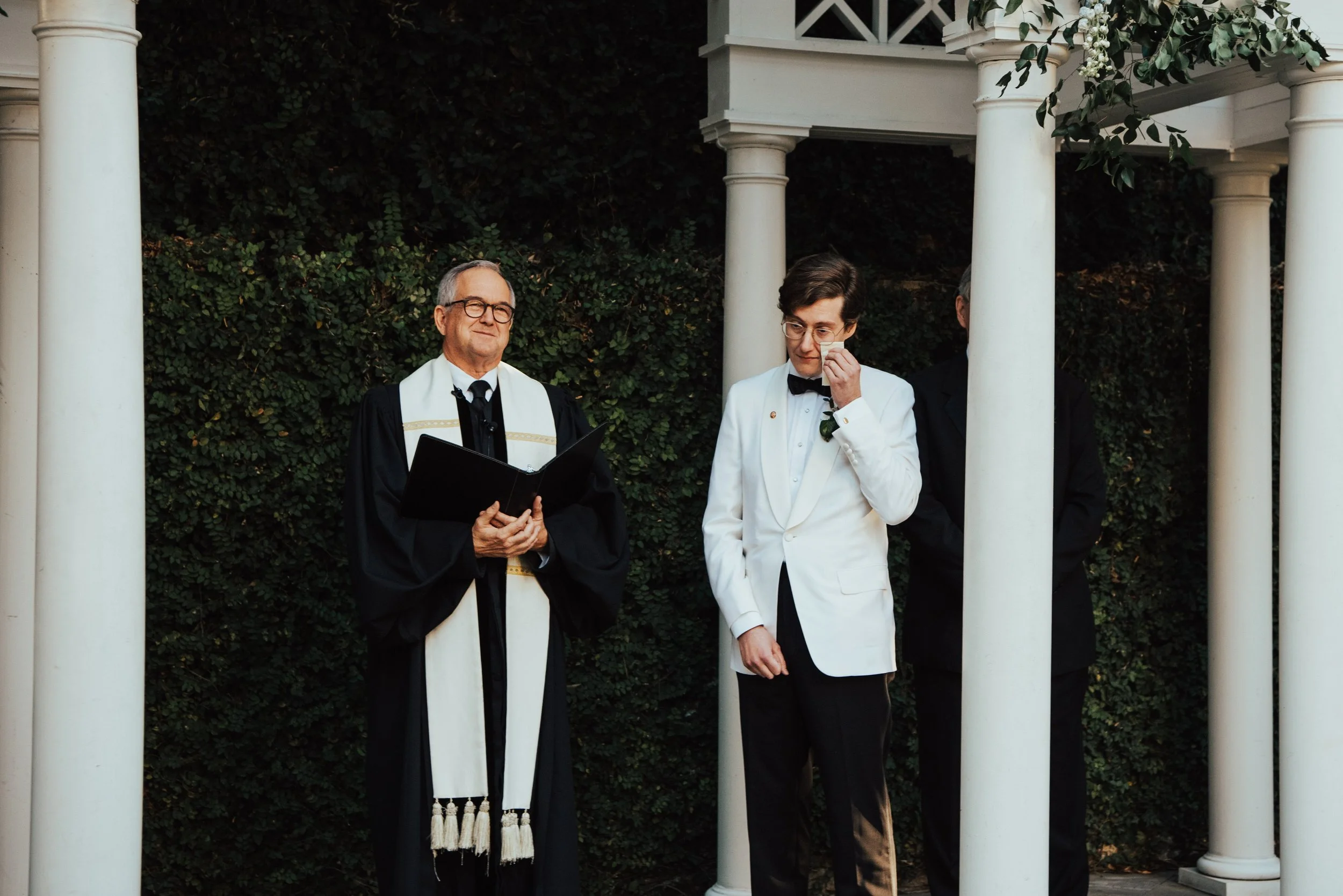 Groom in white tuxedo jacket wiping away a tear with the minister smiles beside him