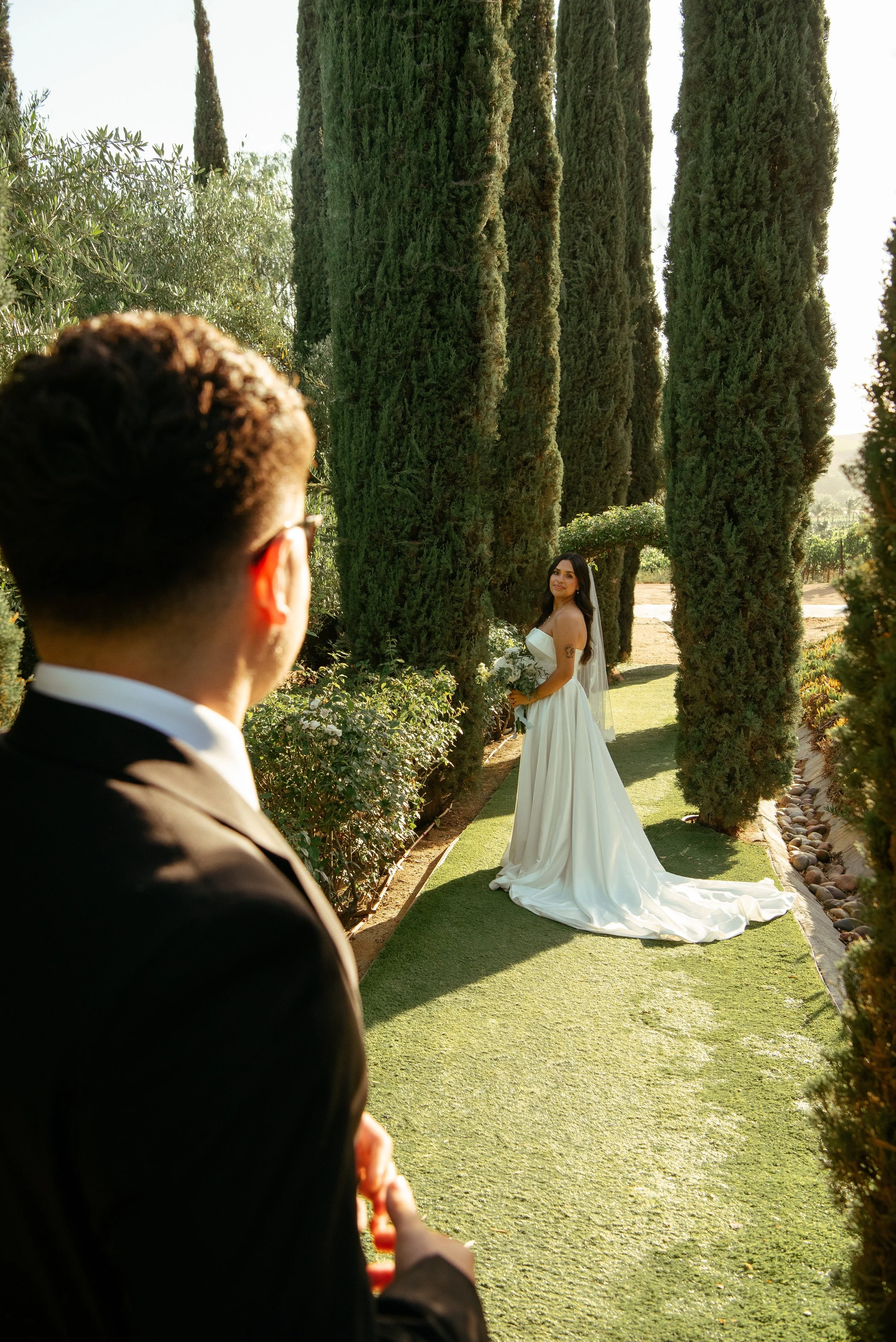 Shot over grooms shoulder as he look to his bride farther down the path