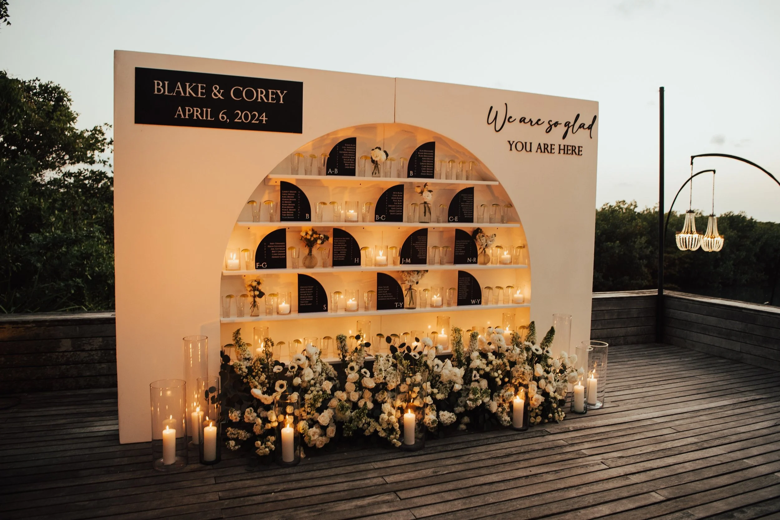 shots of giant welcome sign for wedding reception. Candles and white florals all along the bottom with little black signs with guest seating on shelfs
