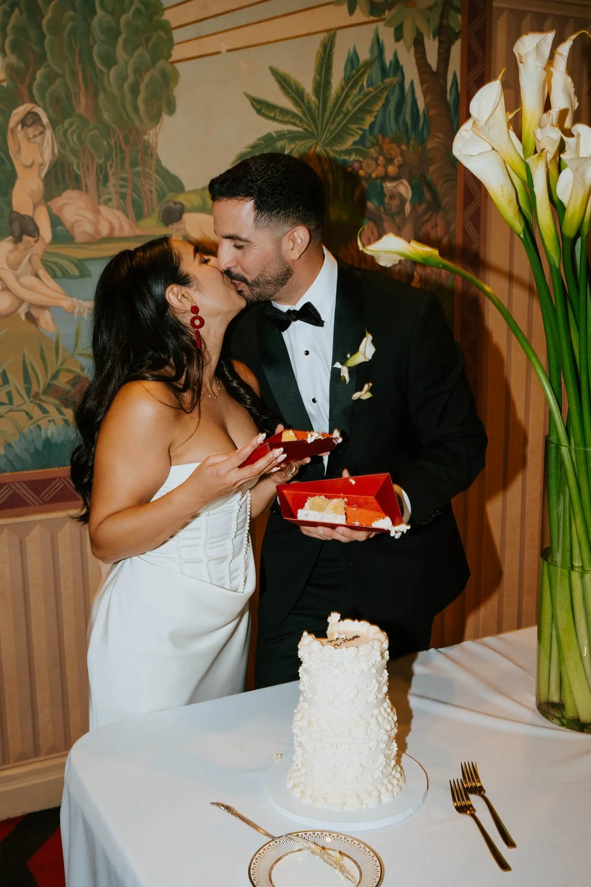Bride and groom holding cake and kissing in from of a mural