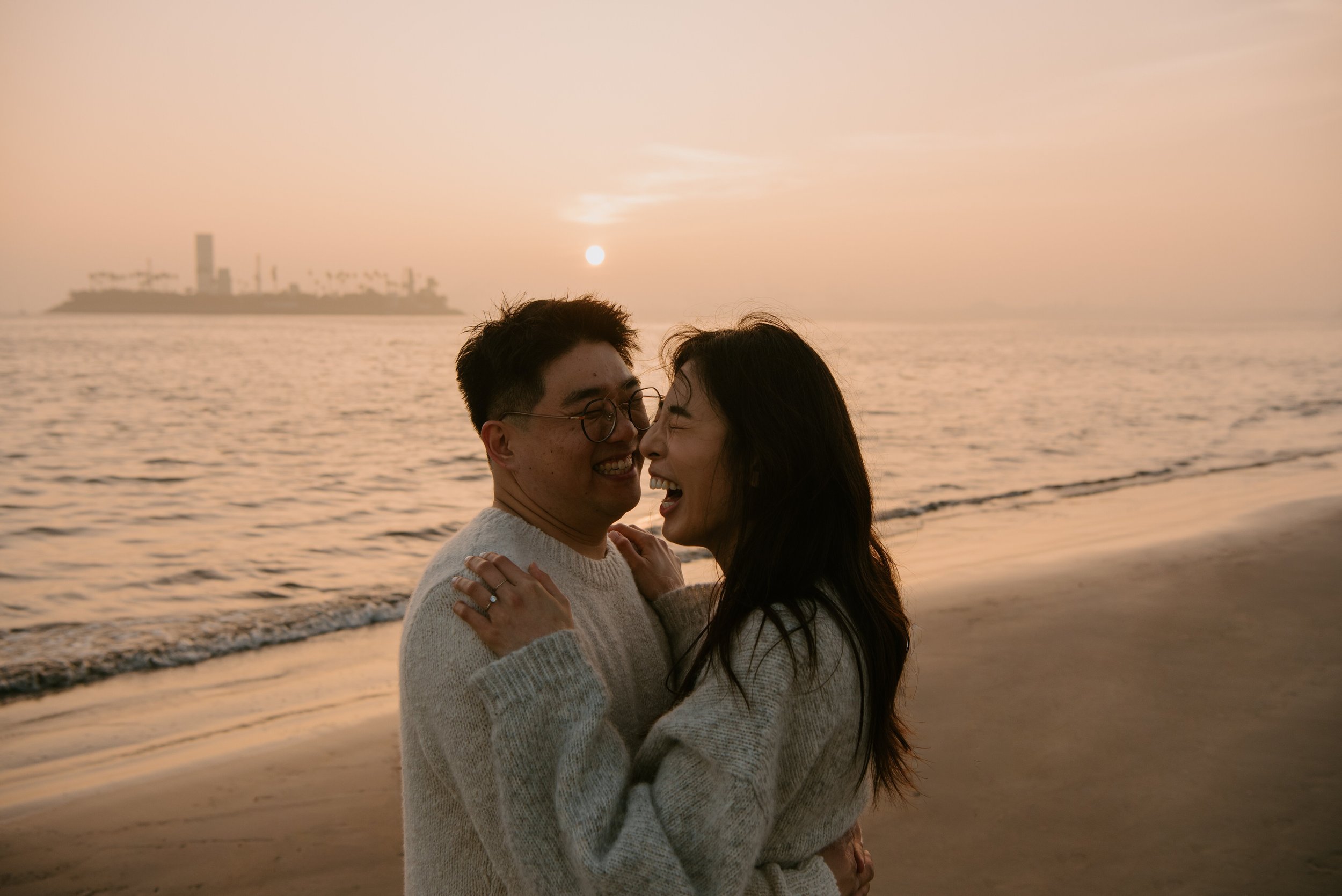 Couple embracing and laughing on the beach with the sun setting behind them
