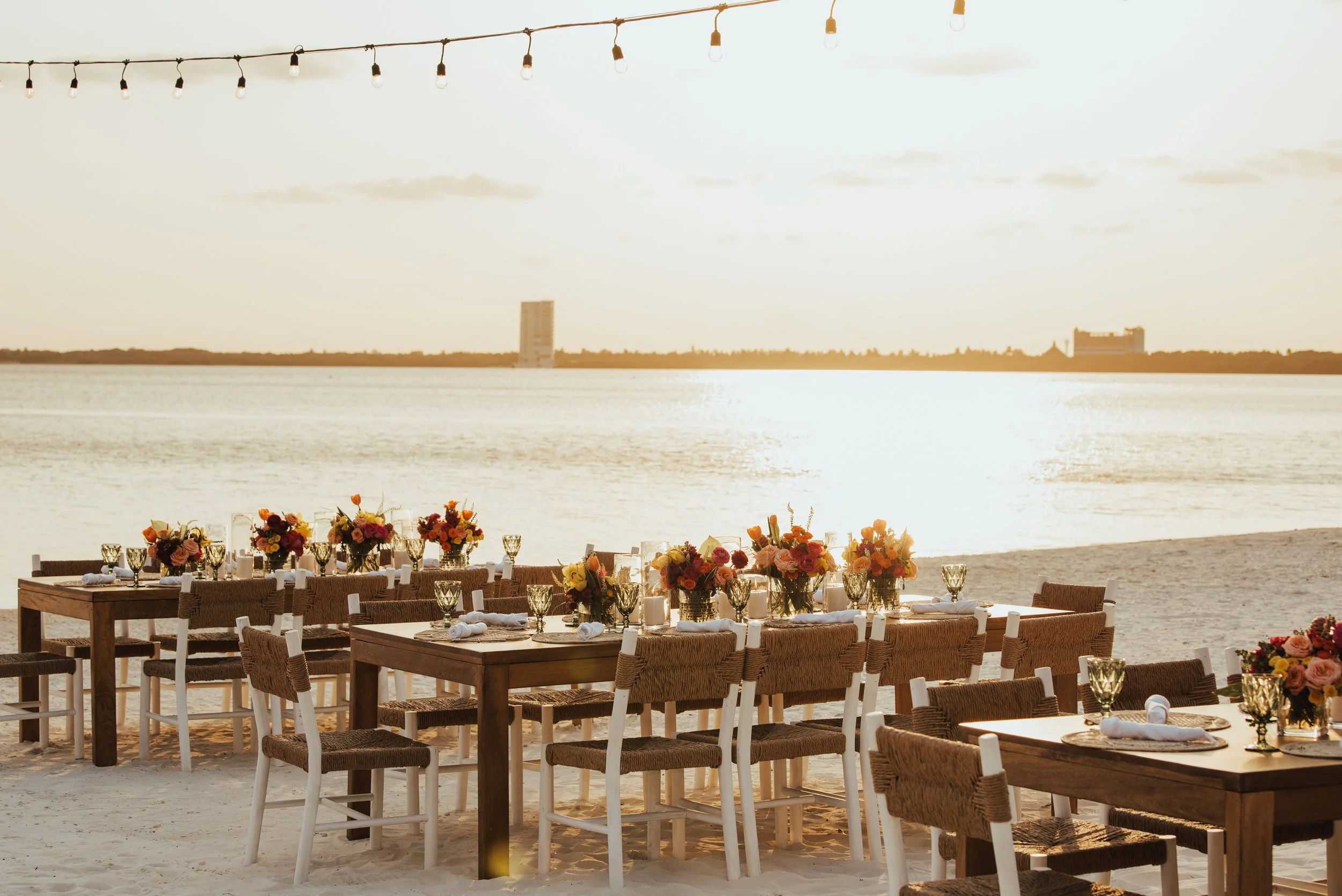 side view of 3 tables set up for a wedding welcome part on the beach with the sun setting behind them