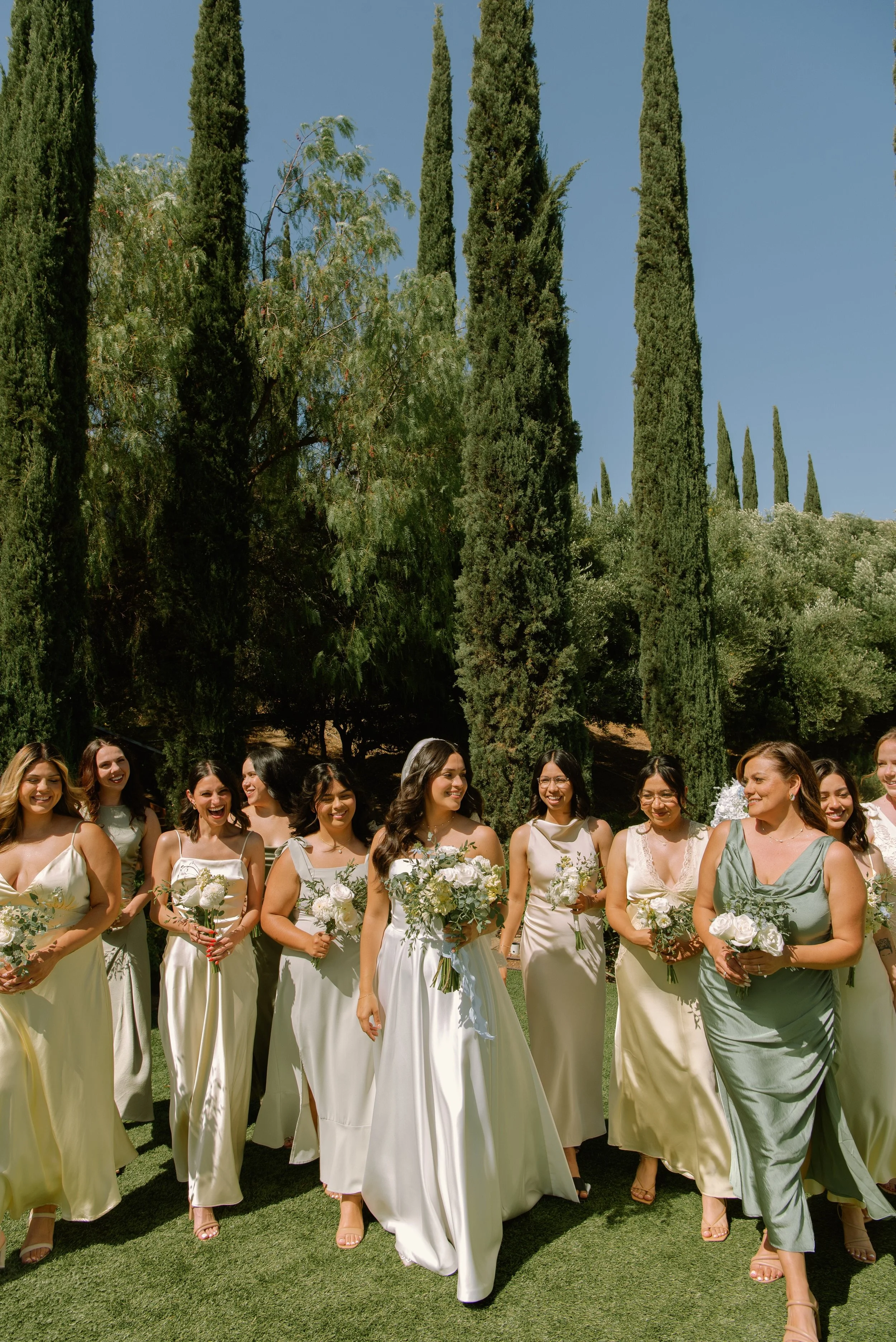 bride and bridesmaids walking toward the camera with tall Italian cypress tress in the background. Bridesmaids are wearing yellow, green, and gold dresses