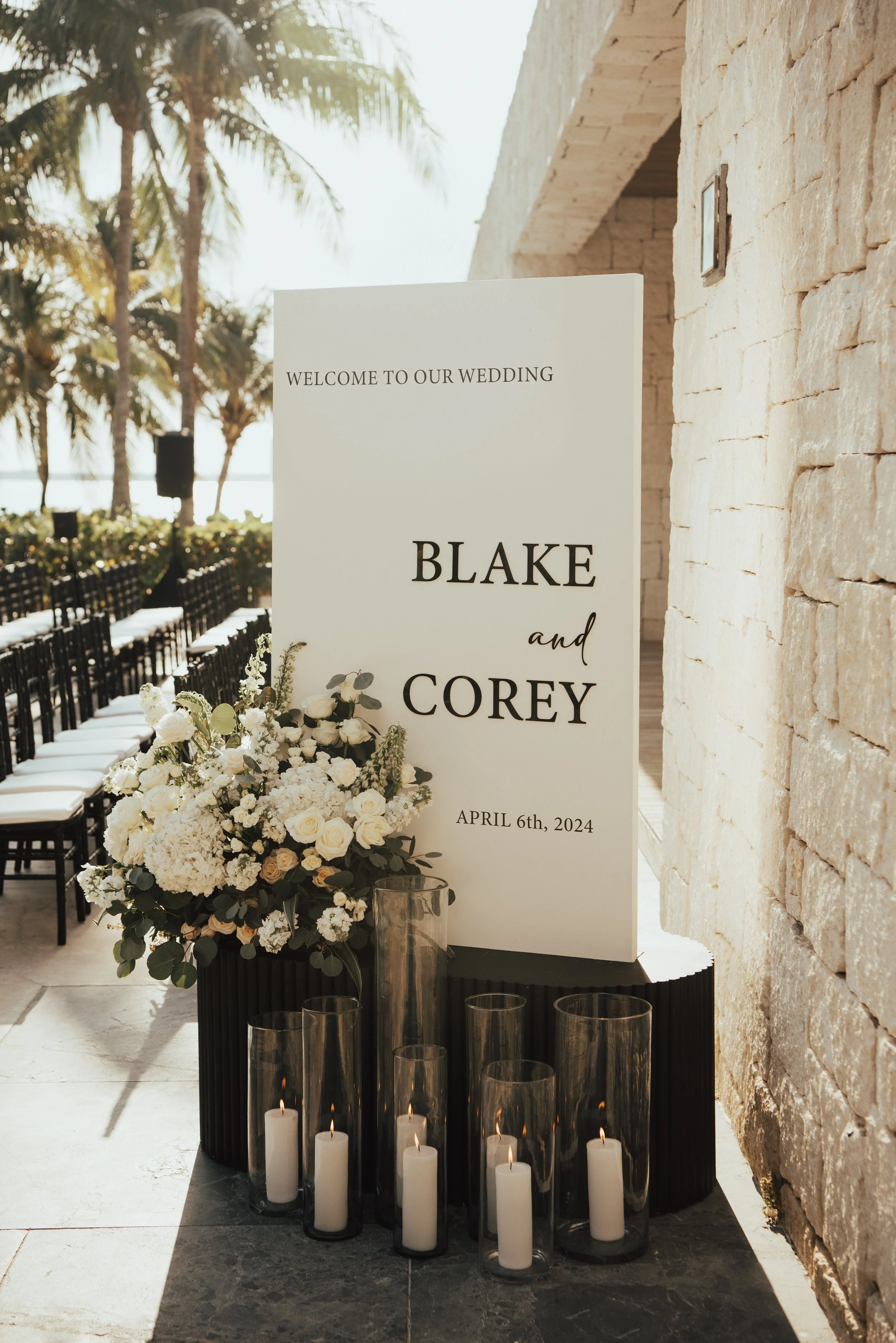 Wedding welcome sign with white florals and candles around it. Palm trees seen in the background.
