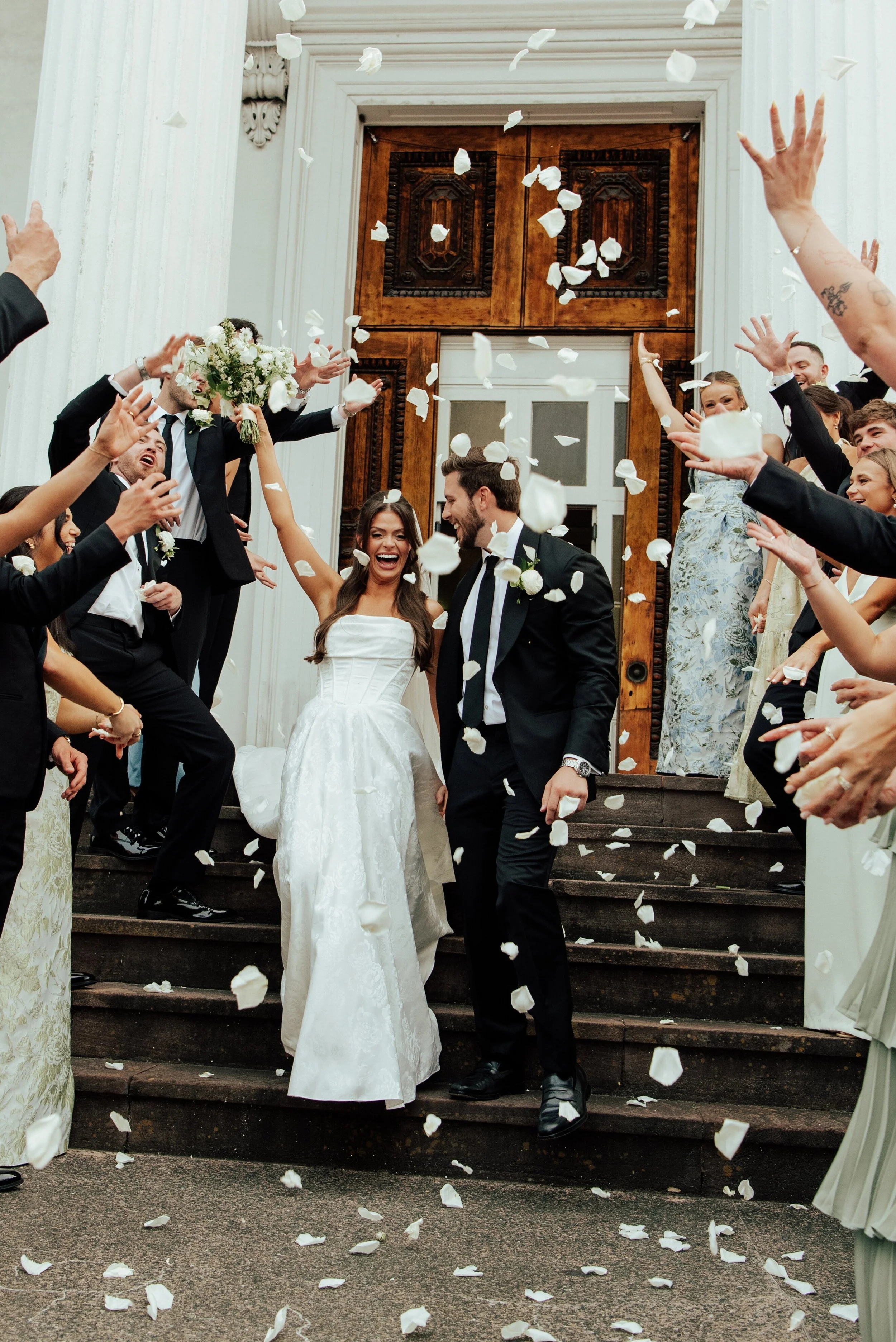 Bride and groom walking down the stairs of a church smiling as their guest throw flower petals in the air