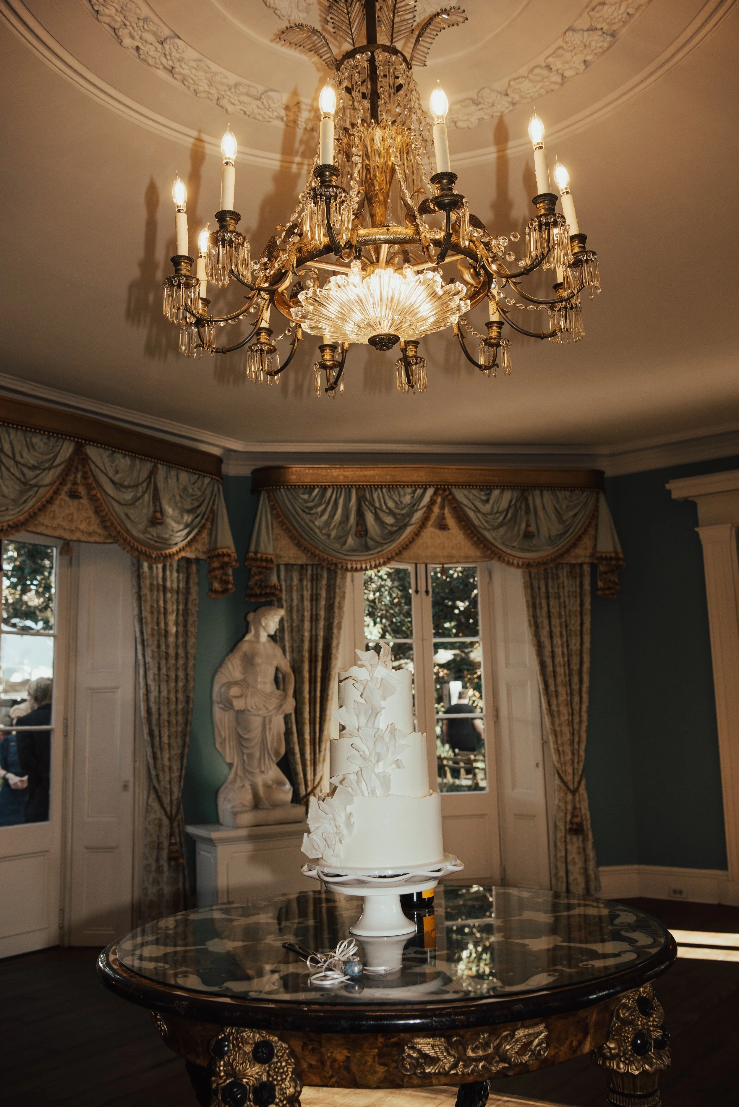 all white wedding cake sitting on round table. Chandelier is hanging from the ceiling and a stone statue is seen in the background