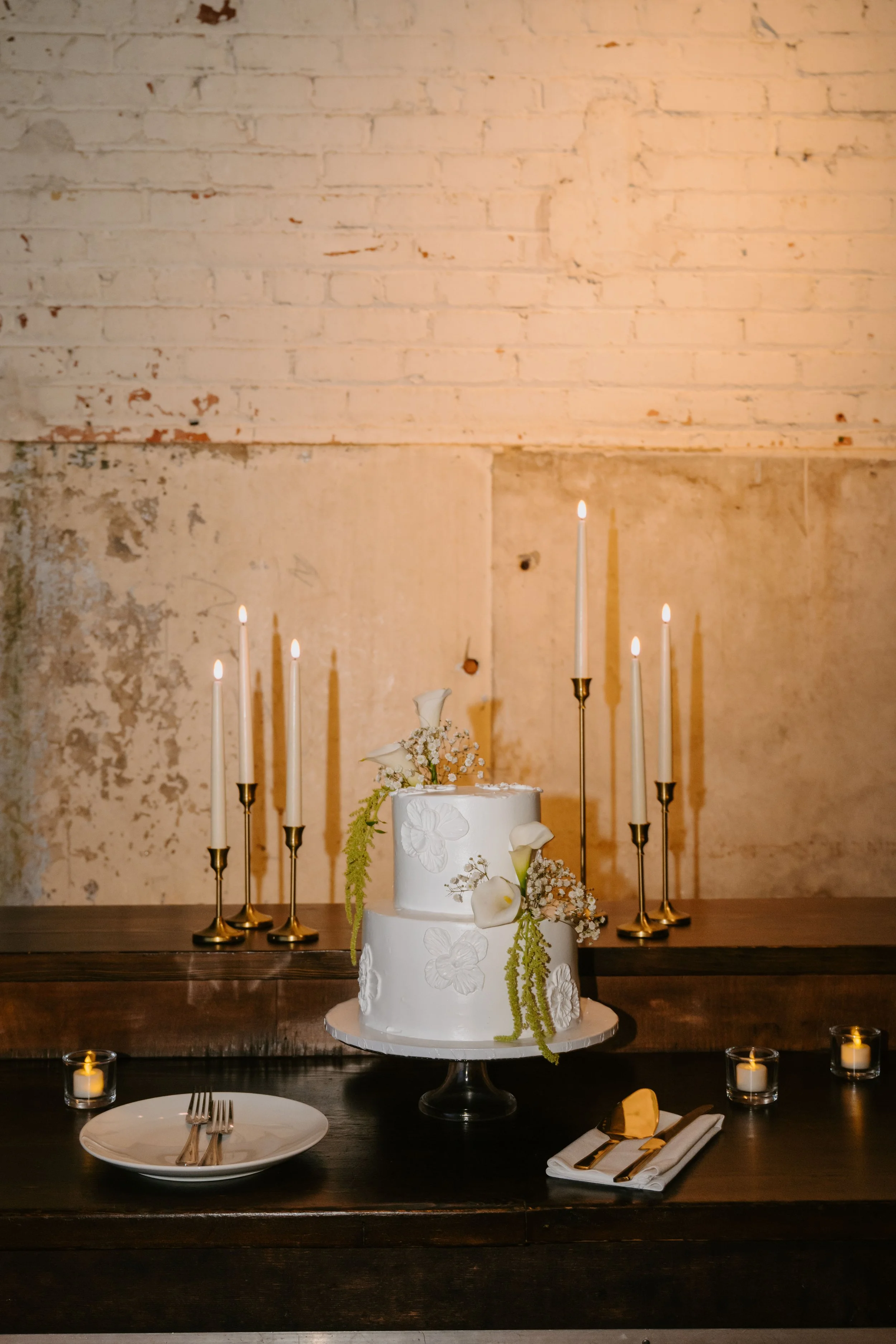 photo of the wedding all white wedding cake with white flowers with some greenery decorating it. Candles are behind the camera
