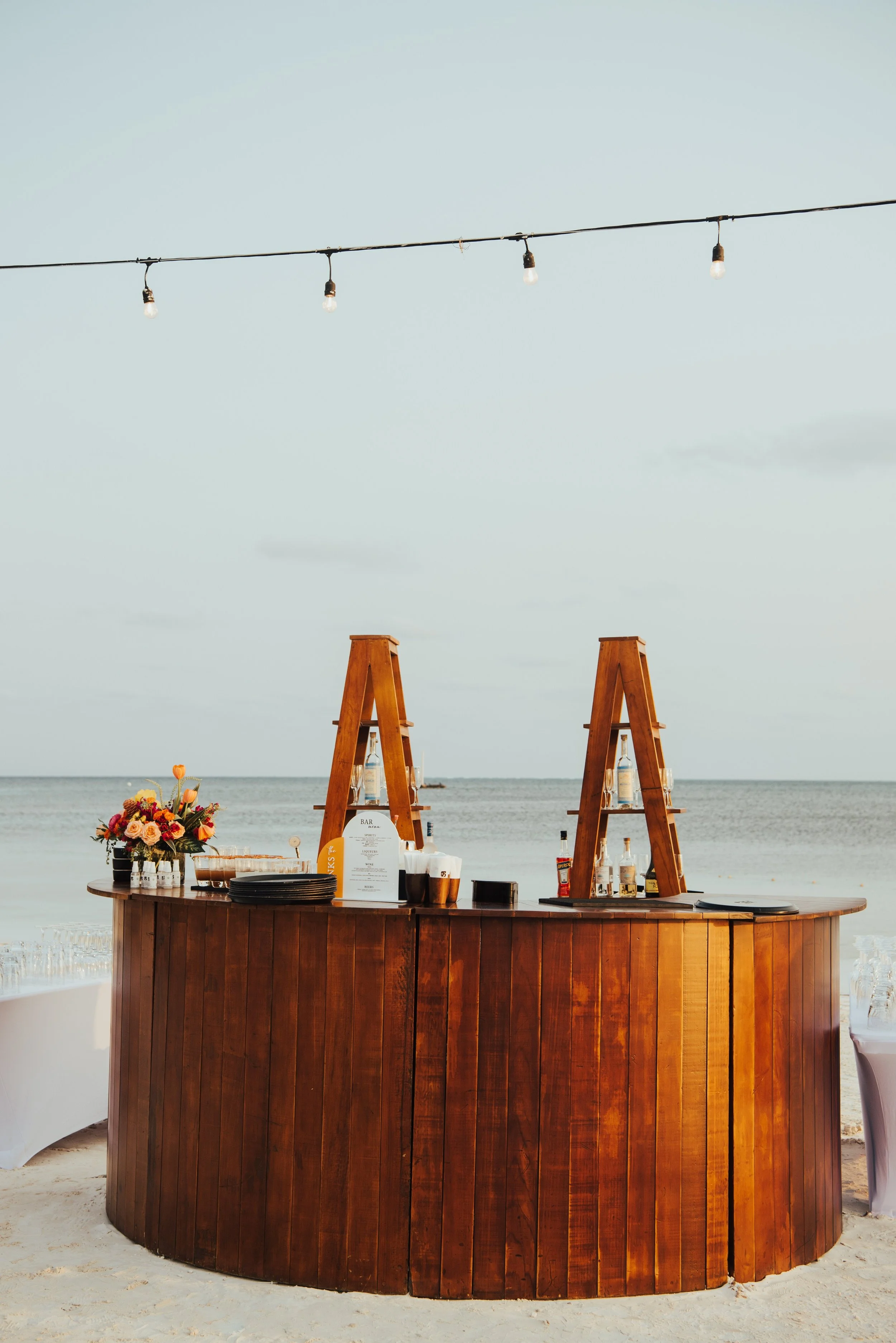 Round wooden bar setup on the beach with blue water & blue sky behind them