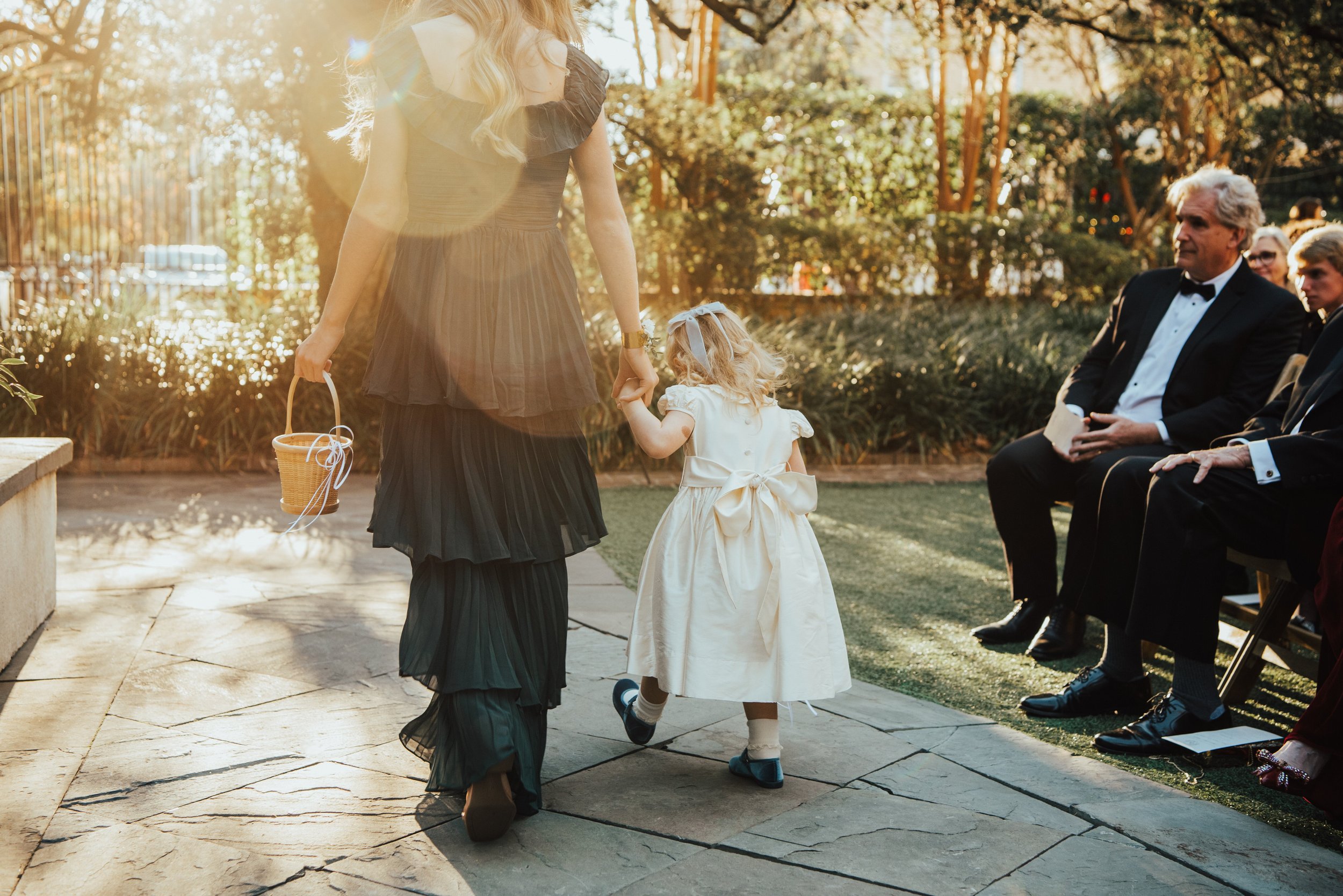 photo of older girl walking away from the camera and holding the hand of the small flower girl. Flower girl has a big bow on the back of her dress.
