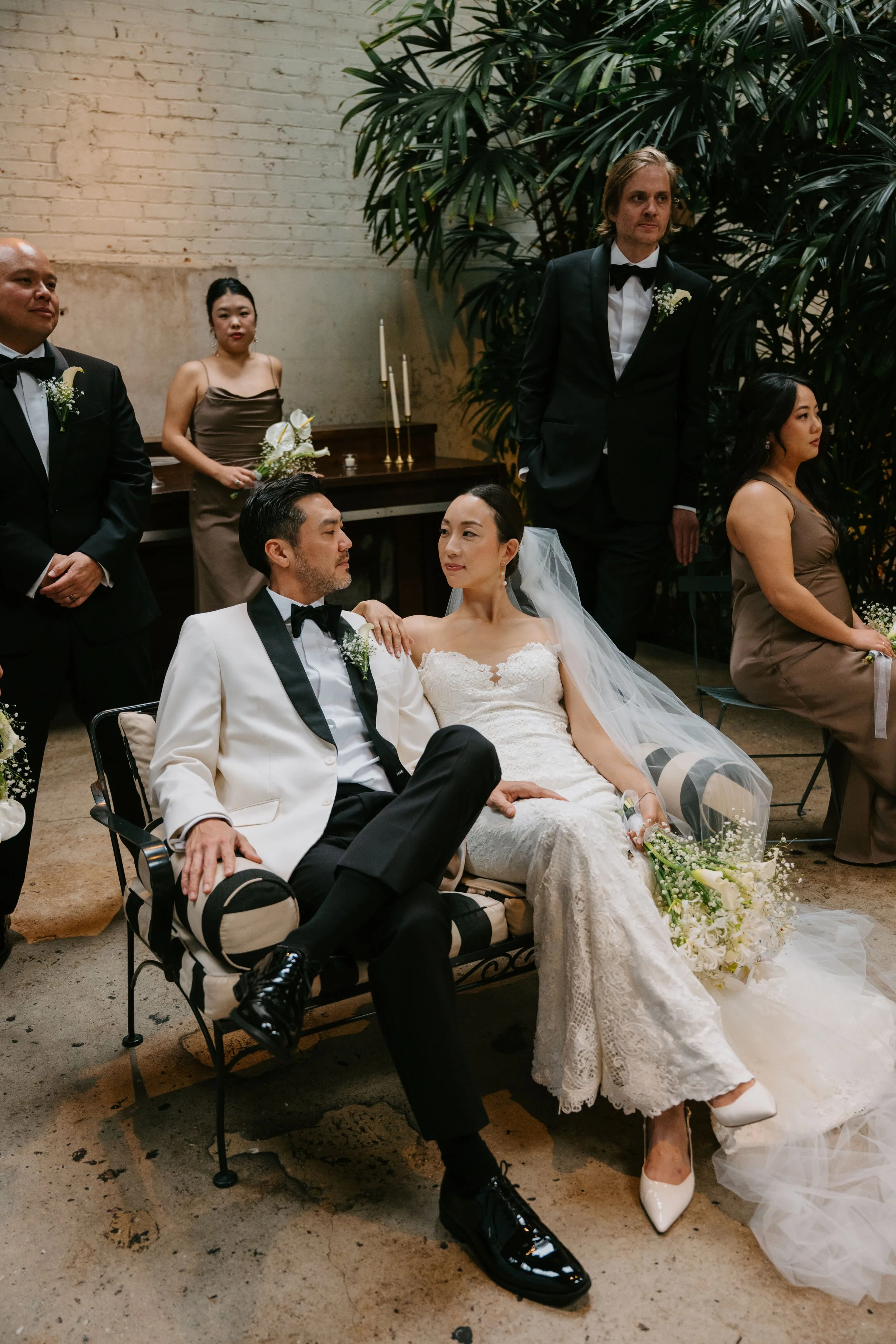 Bride and groom sitting on a couch looking at each other. You can see some of the wedding party behind them