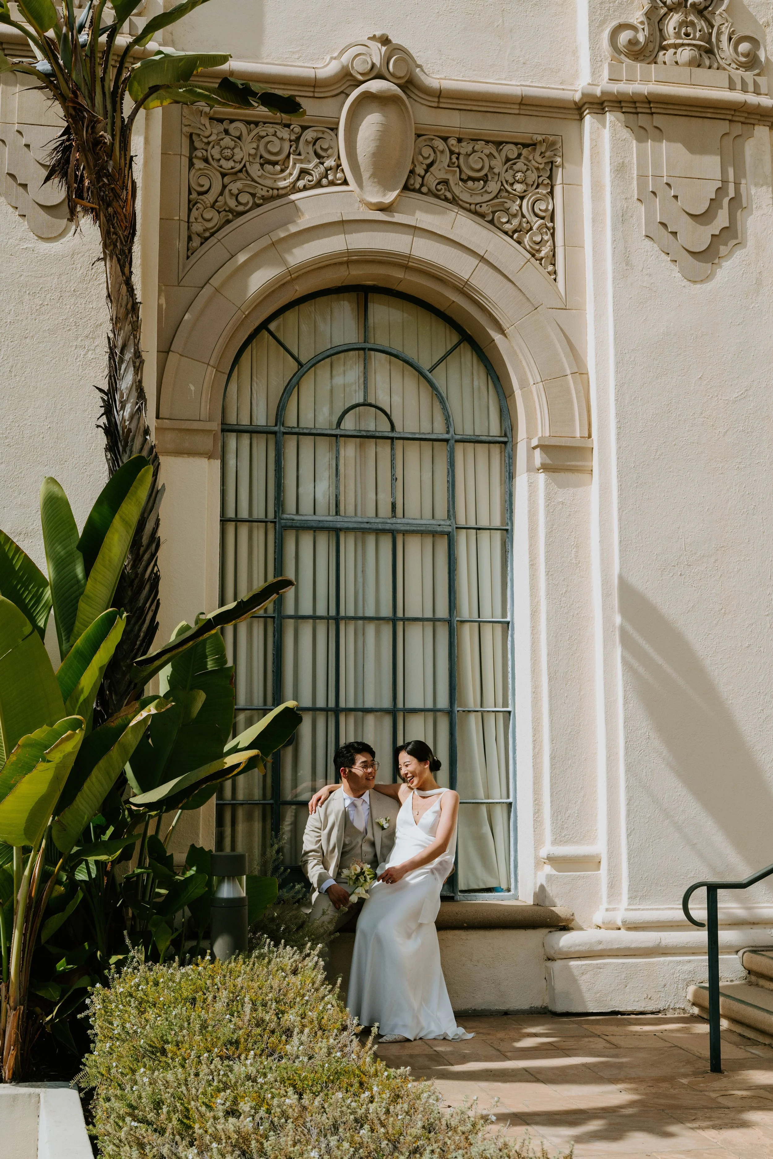 Bride and Groom sitting on the window sill of a large arched window. Couple is laughing together