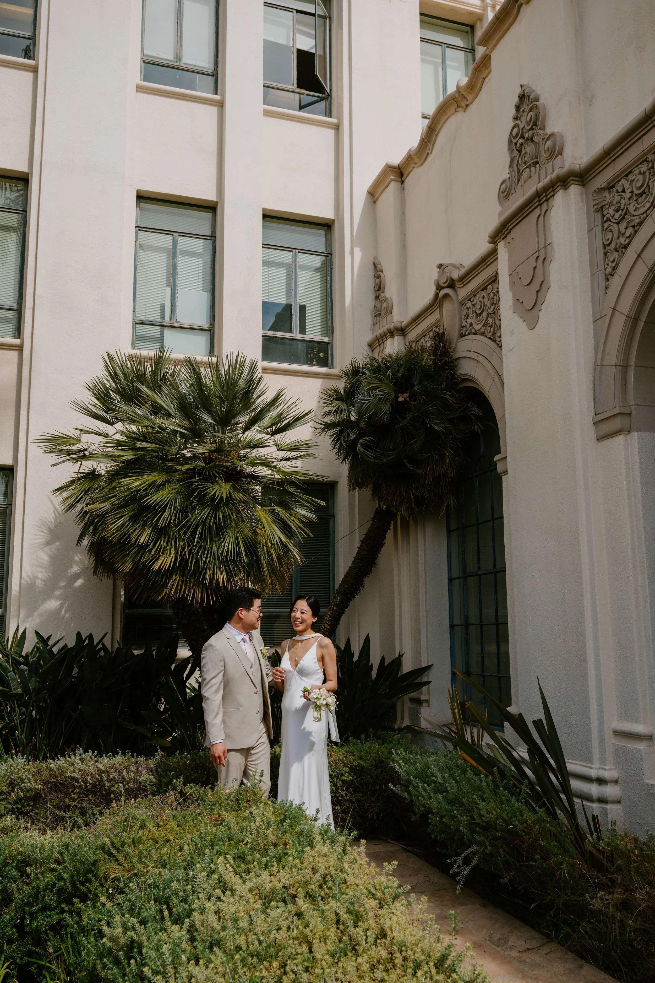 Bride and groom standing and laughing with each other in front of palm trees and city hall