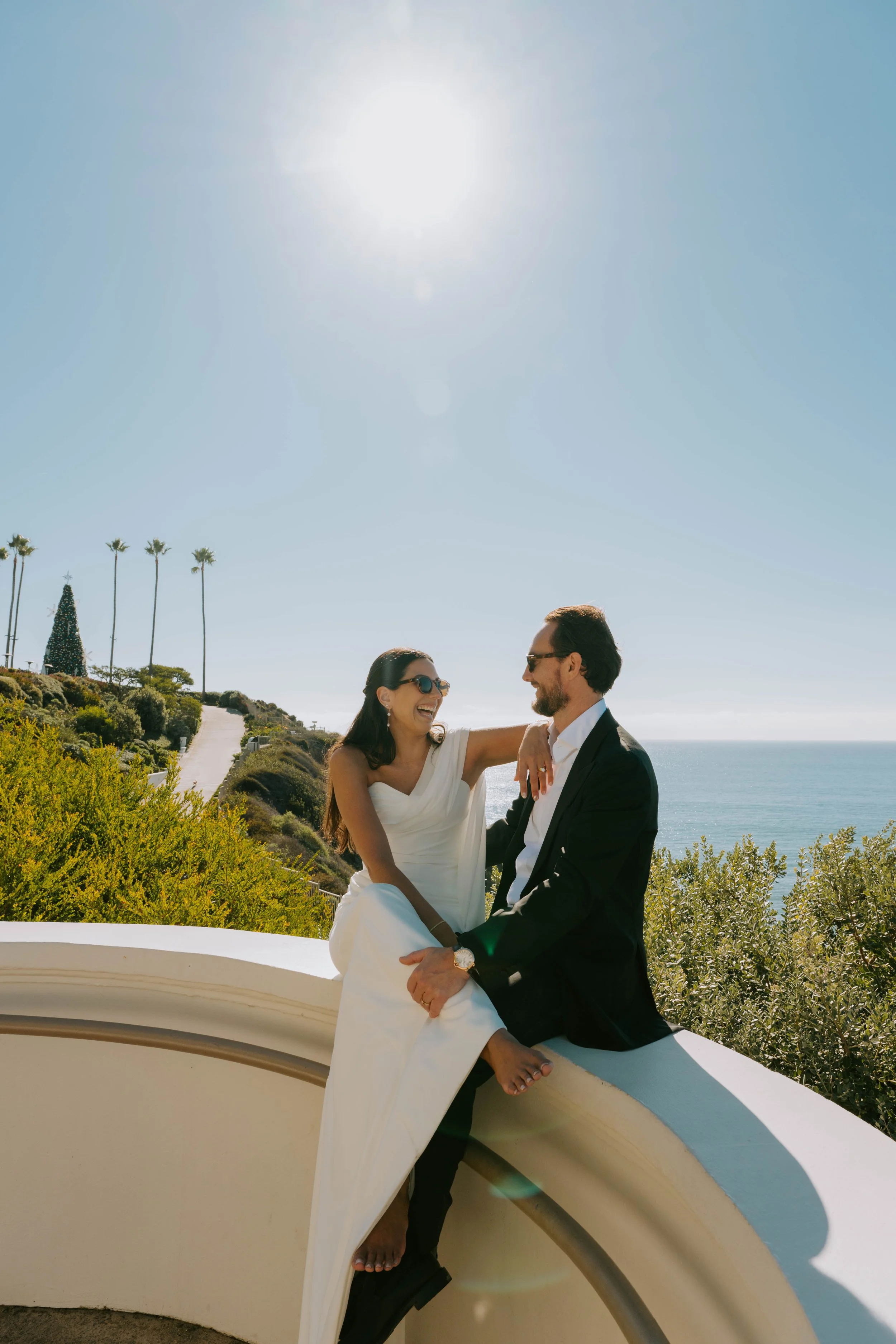 bride and groom sitting and laughing on wall with ocean and blue sky behind them