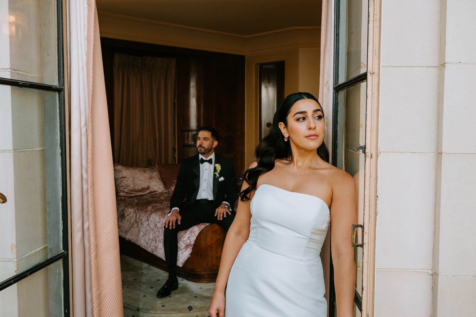 bride leaning against glass door looking of to the side and groom is seen in background sitting on bed looking in opposite direction