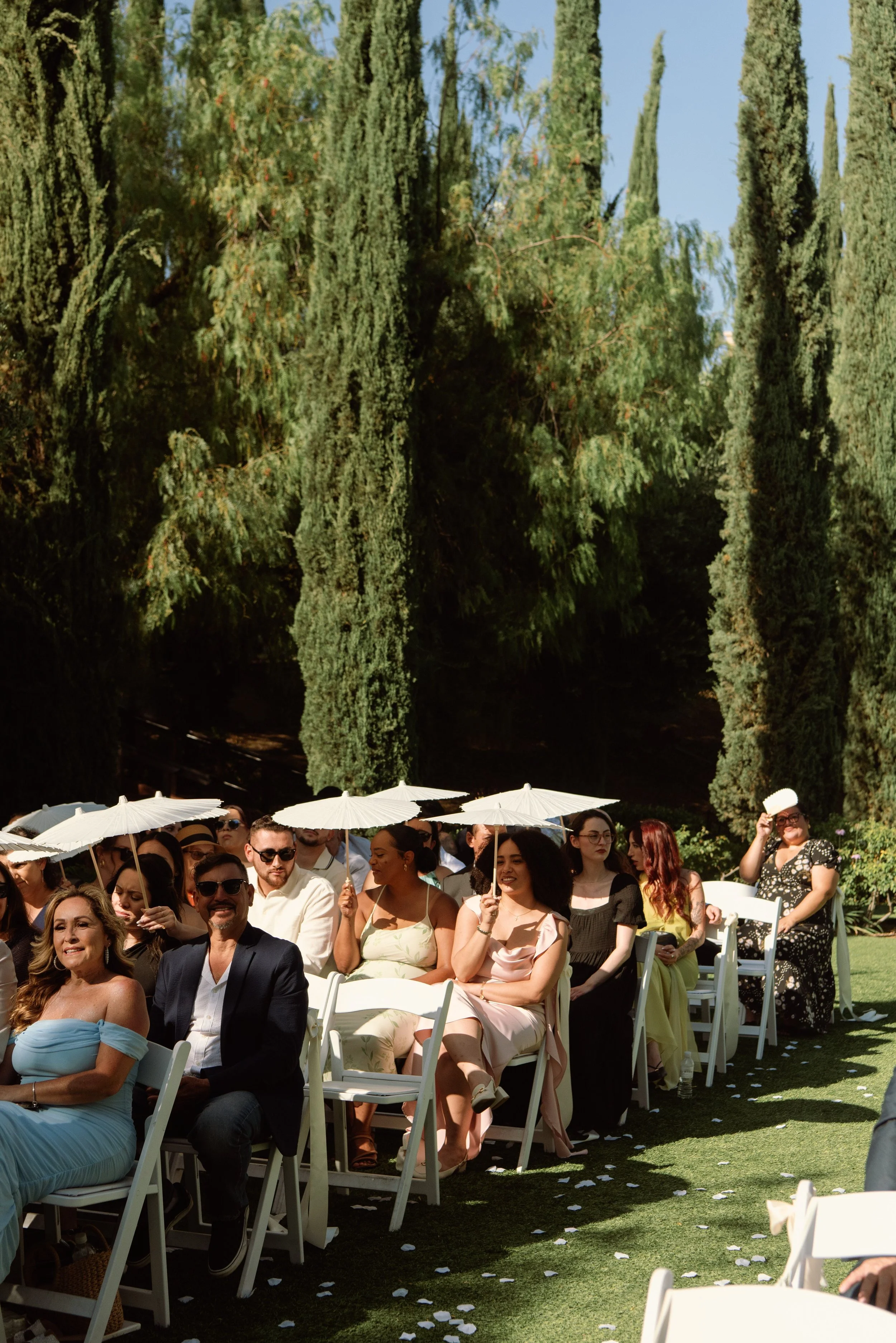 shot of wedding guest sitting at ceremony site. Some guest are holding white umbrellas up to block the sun