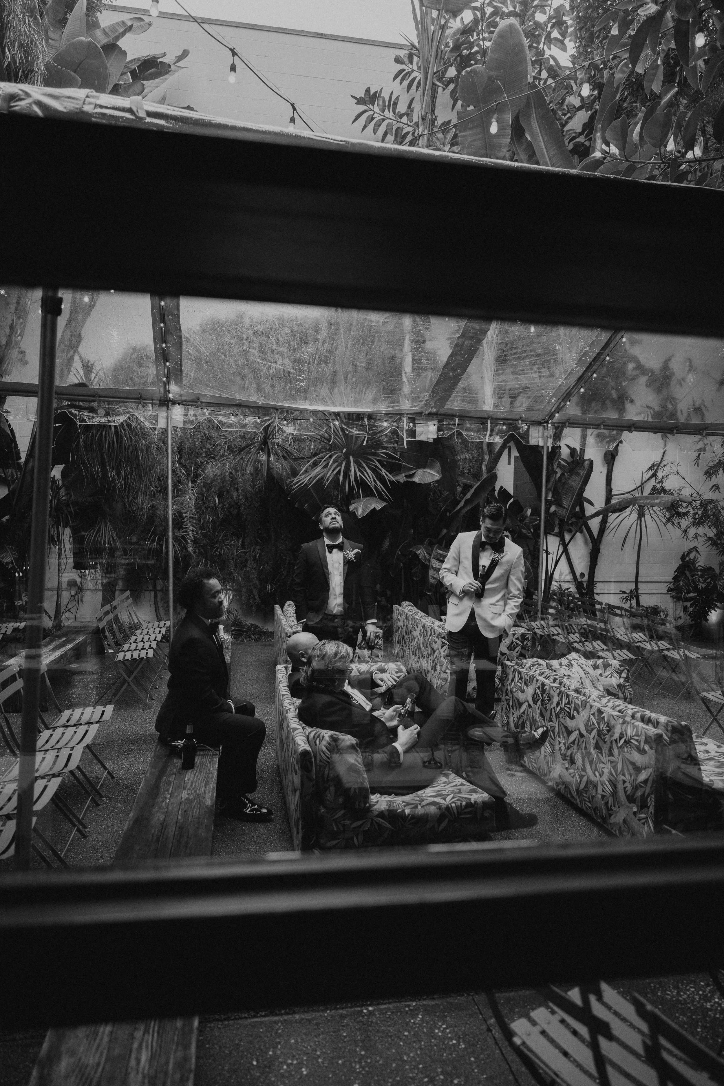 Black and white photo pf groom and groomsmen hanging out. Some sitting some standing. Photo is shot through a window.