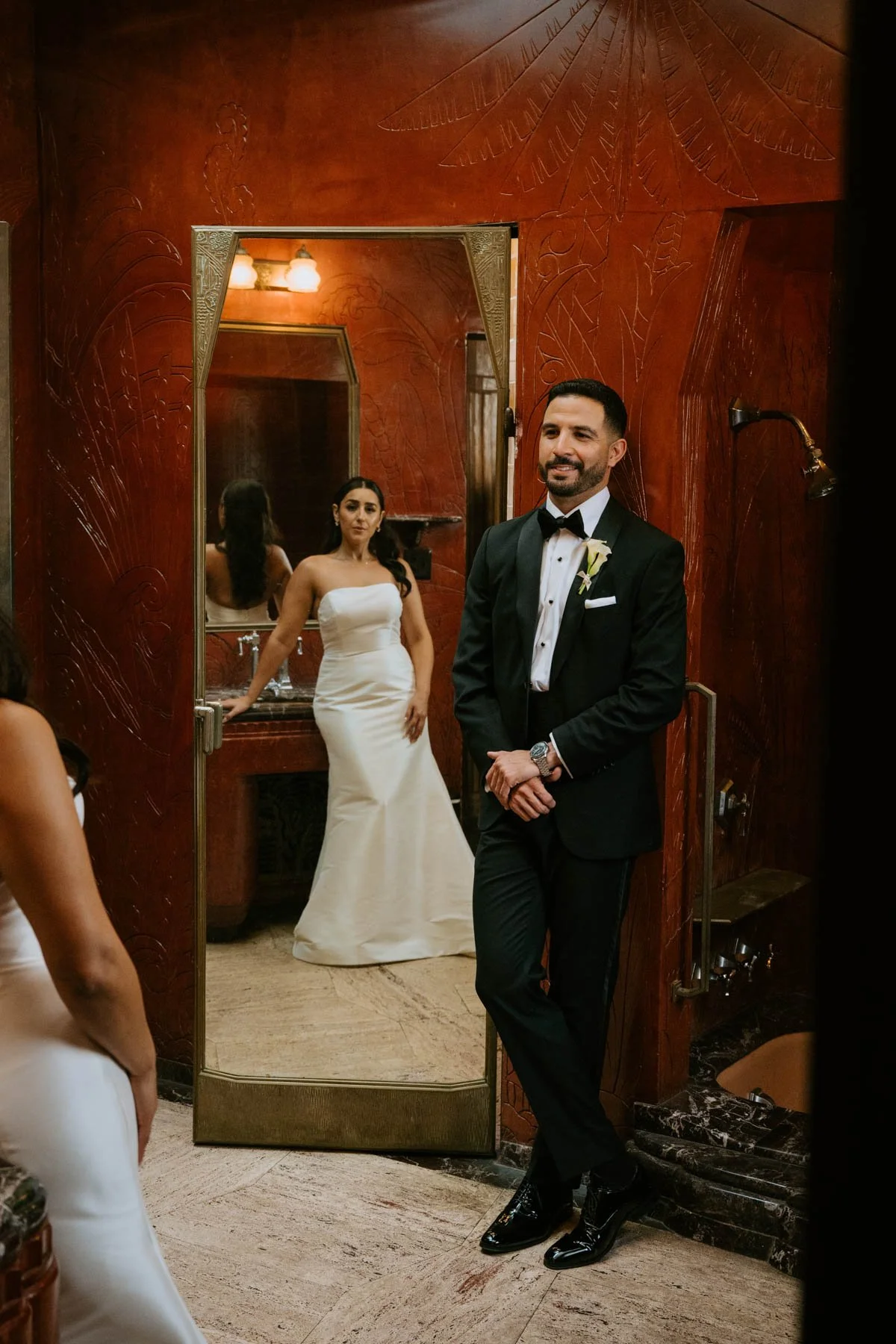 Bride and groom in red bathroom. Groom is leaning against a wall facing the camera but looking at his bride. bride is opposite of him but can be seen in the mirror door next to groom