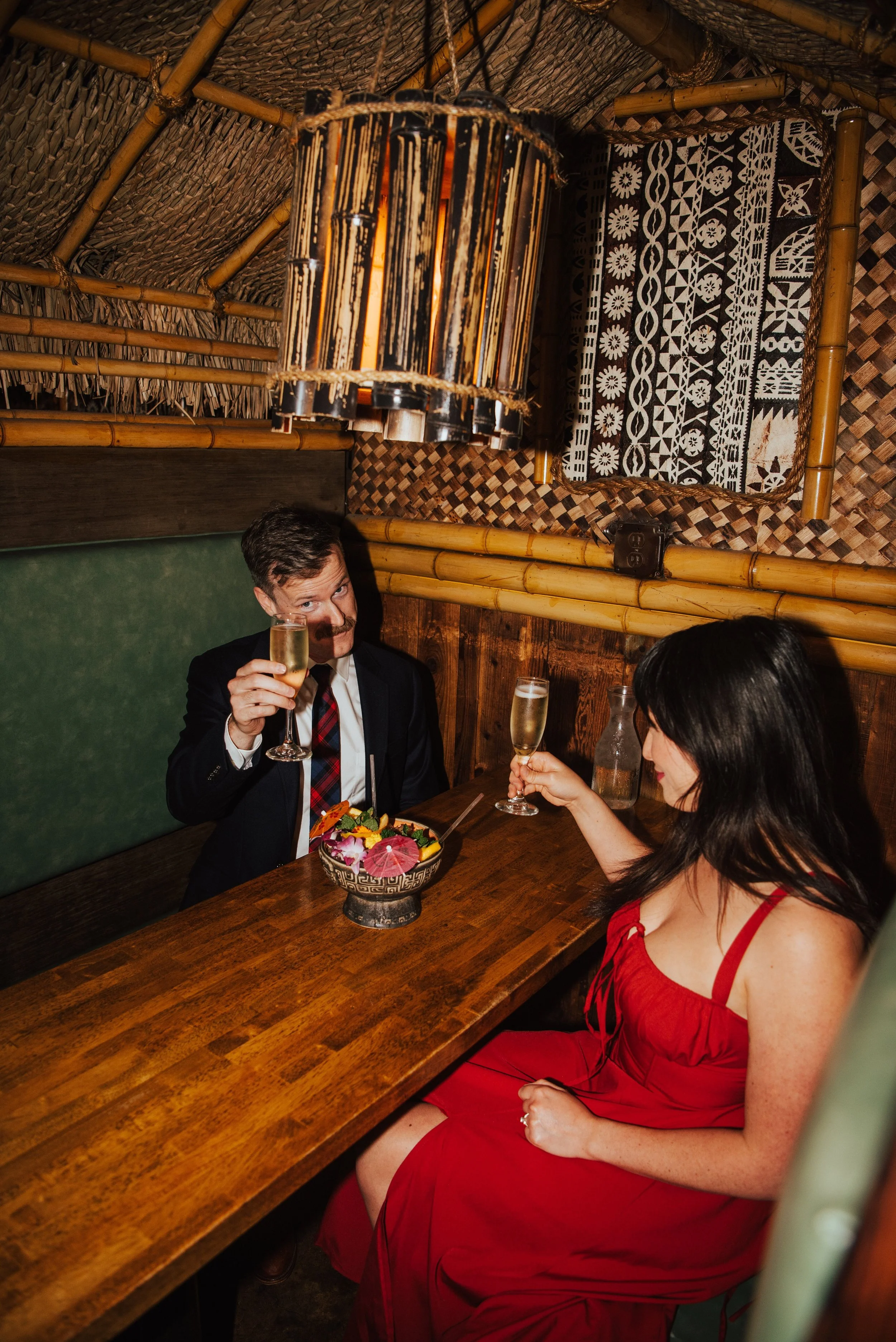 Couple sitting across from each other at a table raising a glass. Guys is looking at the camera as he raises his glass.