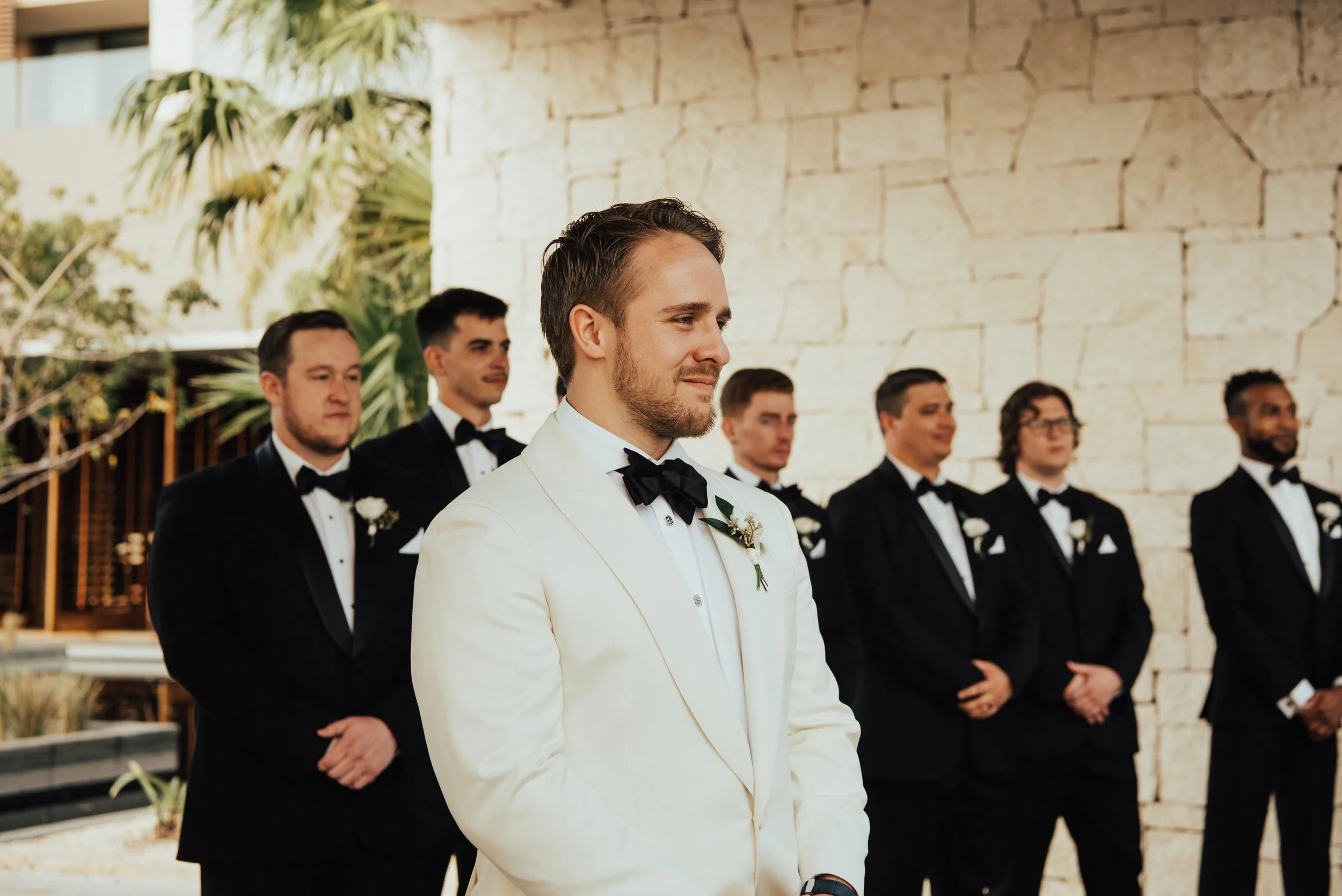 Groom in white suit jacket looking towards his bride. Groomsmen are standing in the background in black suits