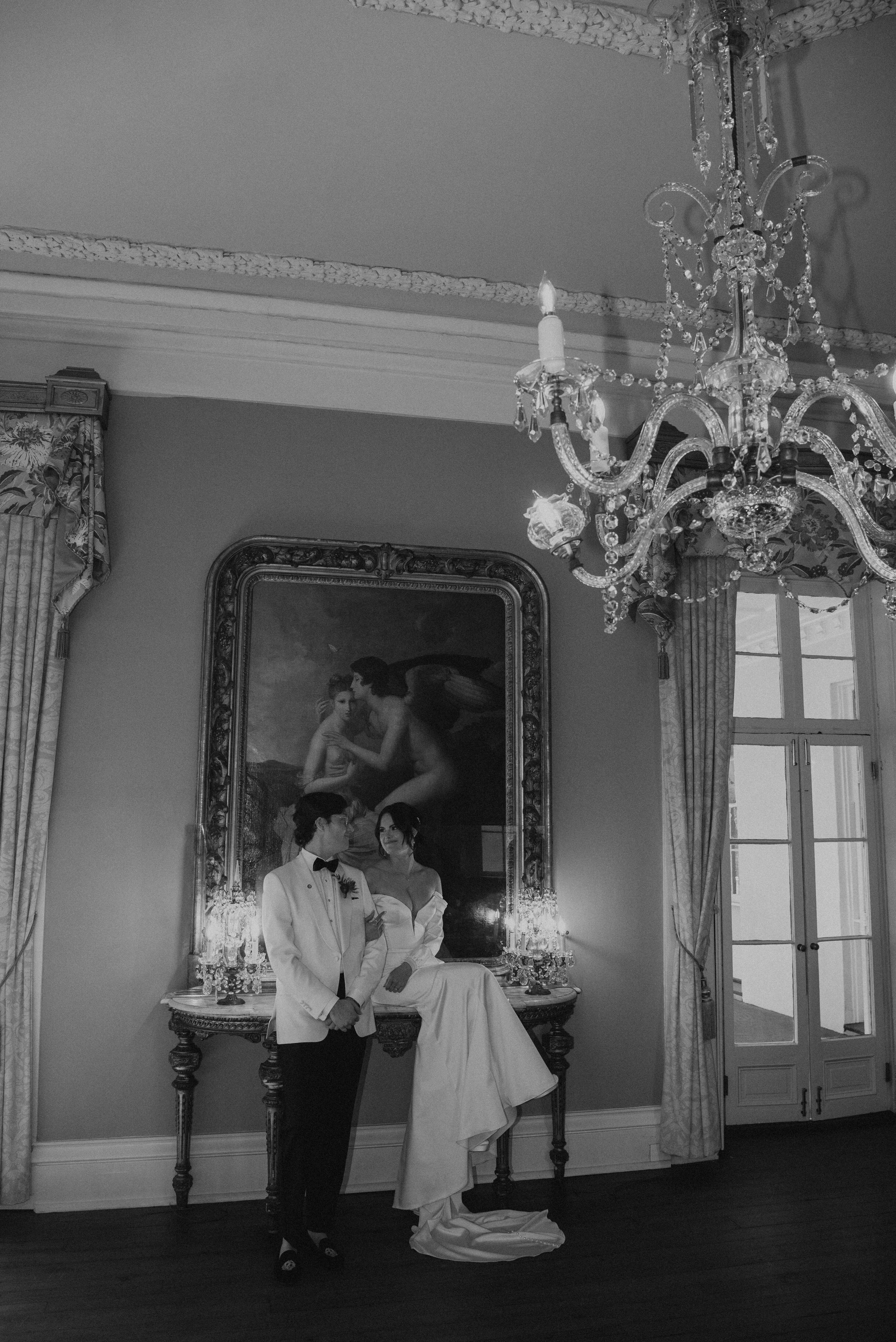 black and white shot of wedding couple in front a large paint with a chandelier in the foreground. Bride is sitting up on a table as she looks at her groom
