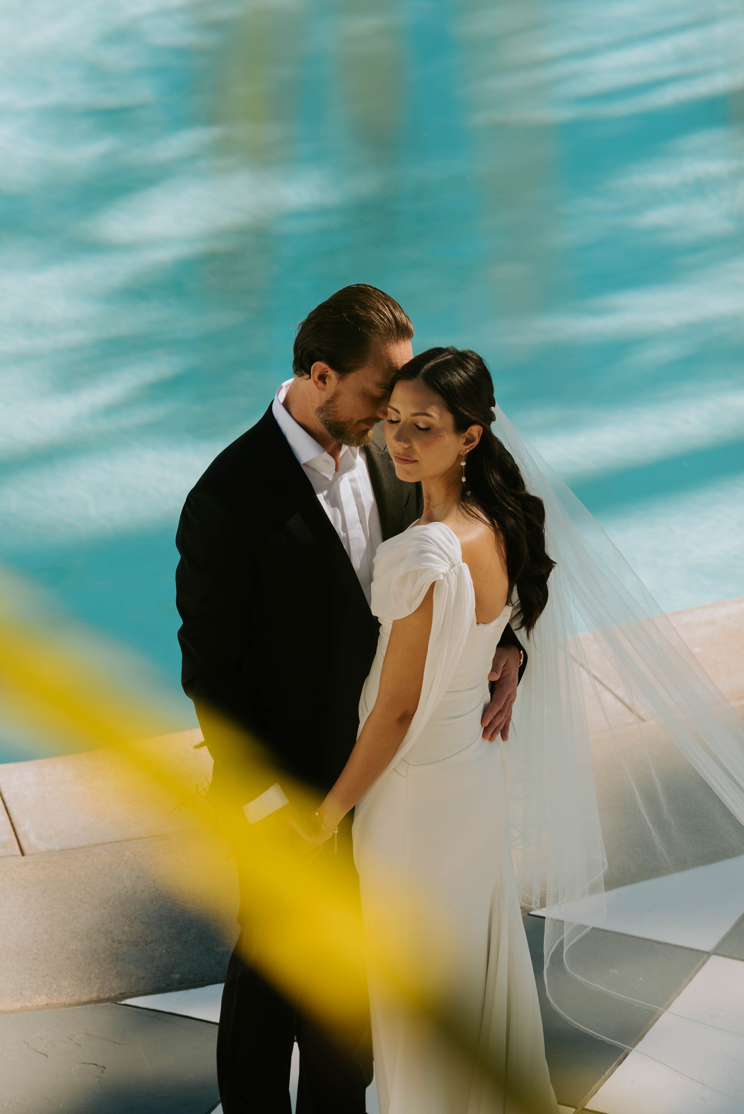Groom resting his head against brides temple as they hug. Pool can be seen in background.