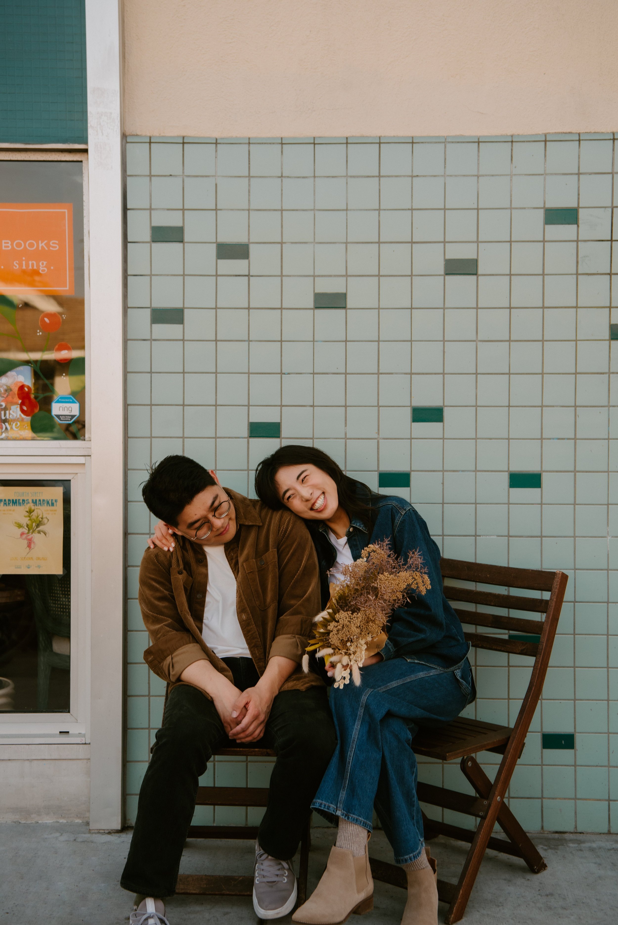 Couple sitting in chairs. Girl leans her head on guys shoulder while she smiles and holds flowers.