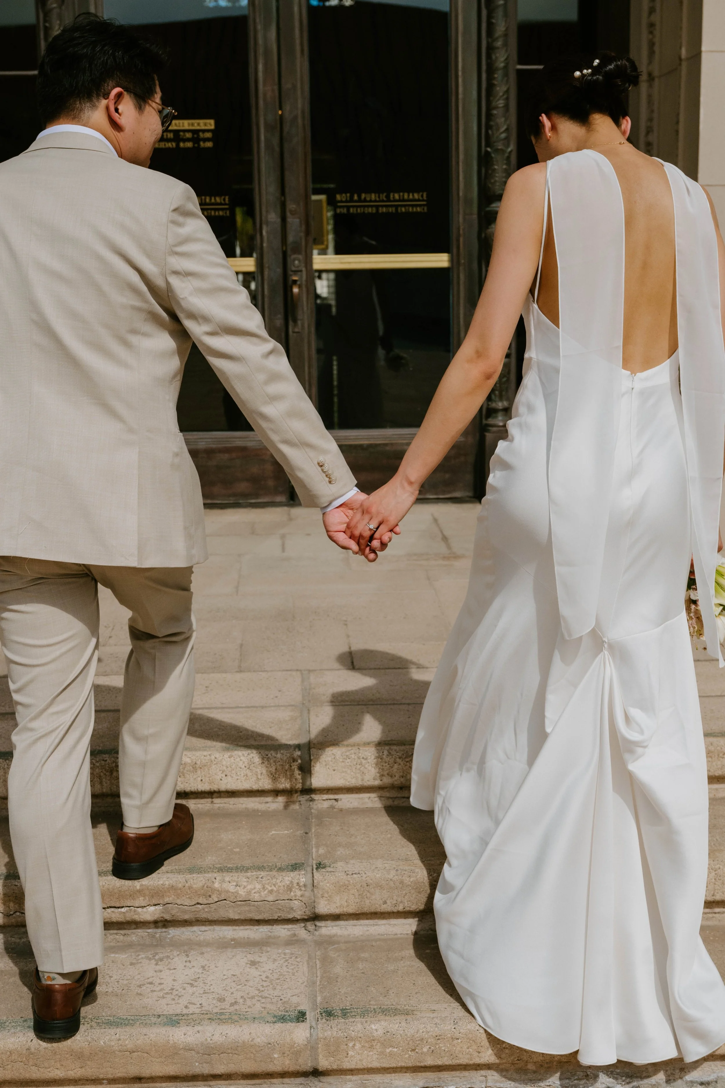 Photo of the back of a bride and groom as they hold hands walking up the steps of Beverly Hills City hall