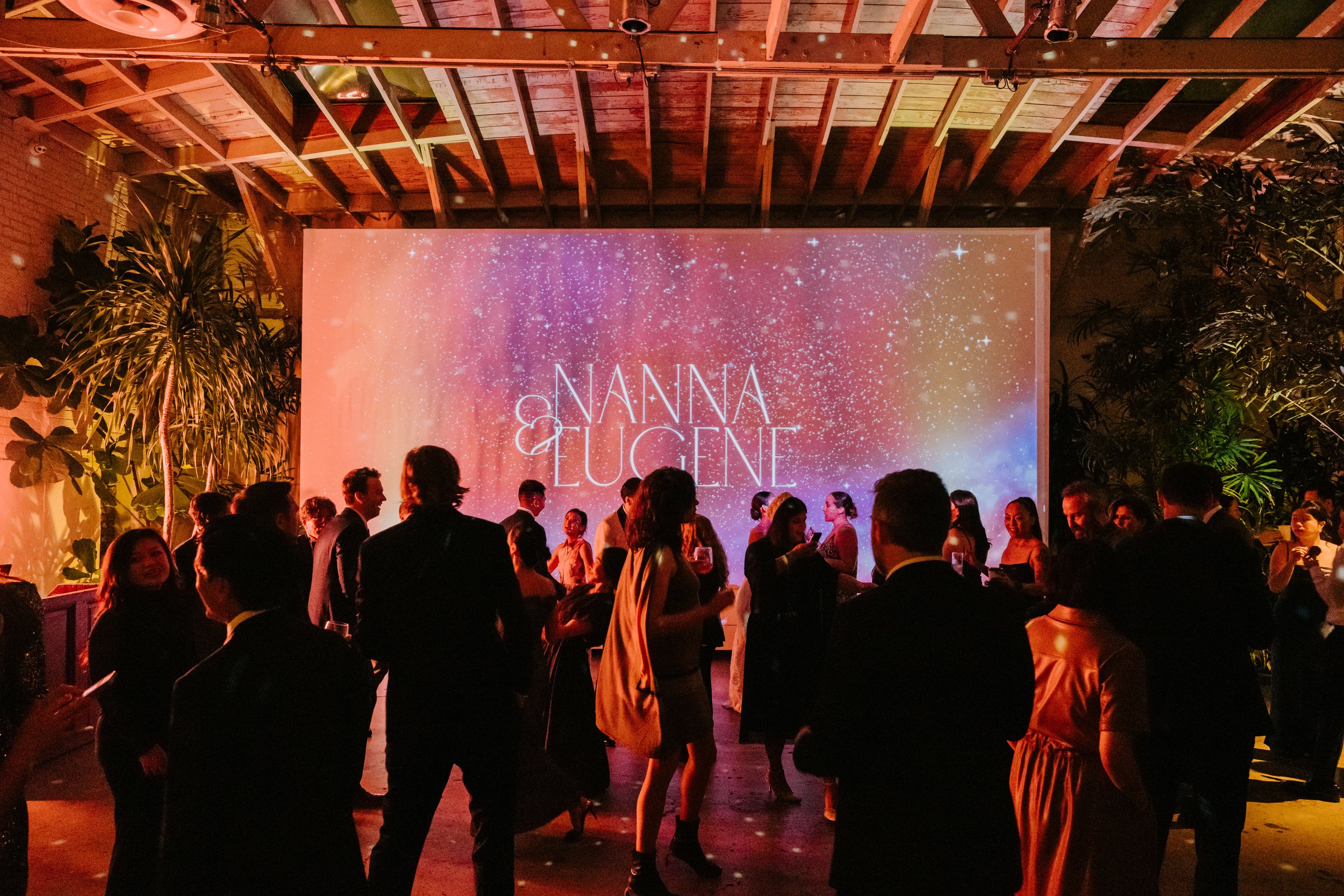 Wide shot of wedding guest dancing with the focus on a large screen in the back with the bride and grooms name on it. Nanna & Eugene