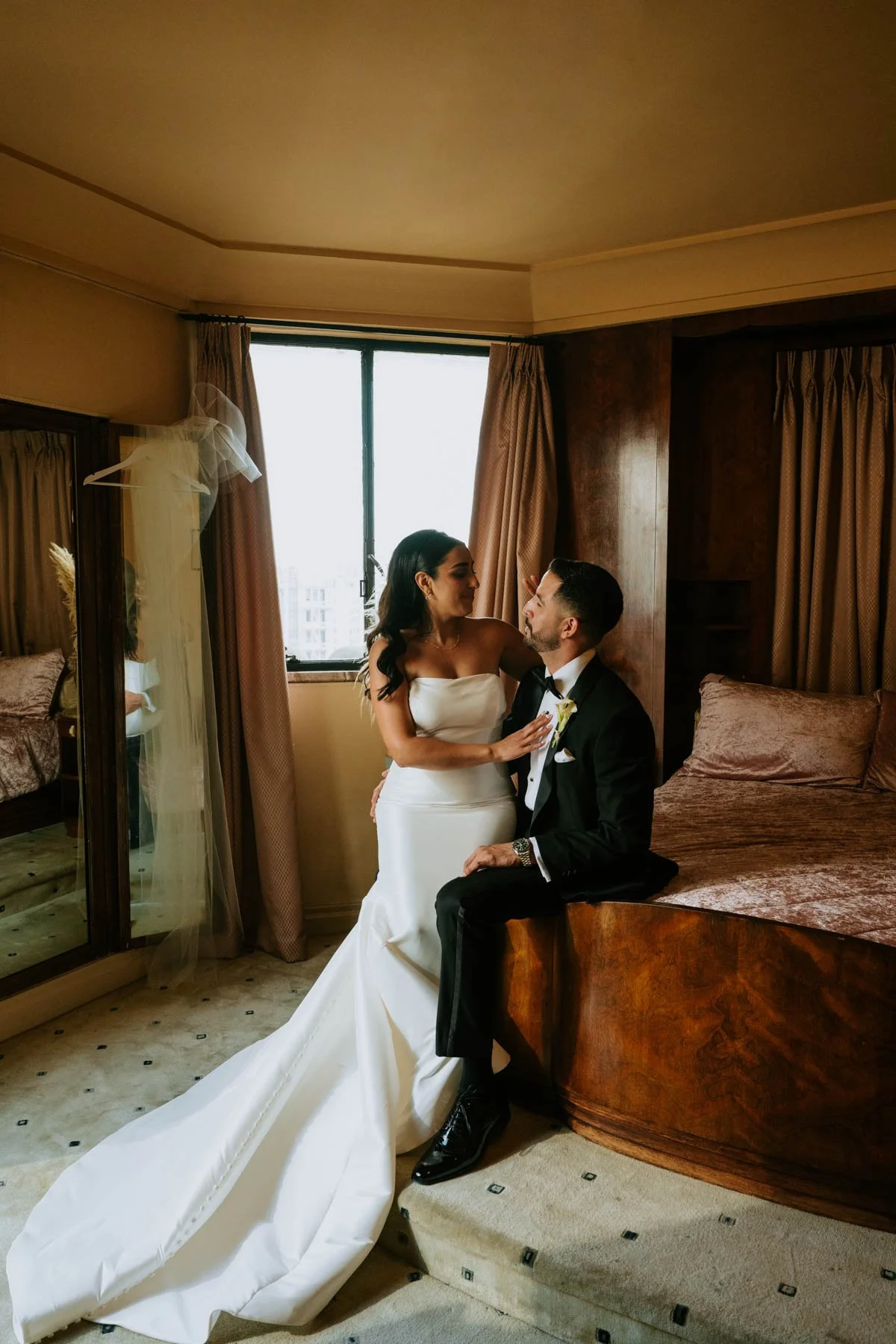 Groom sitting on edge of pink bed with bride sitting on his leg looking at him