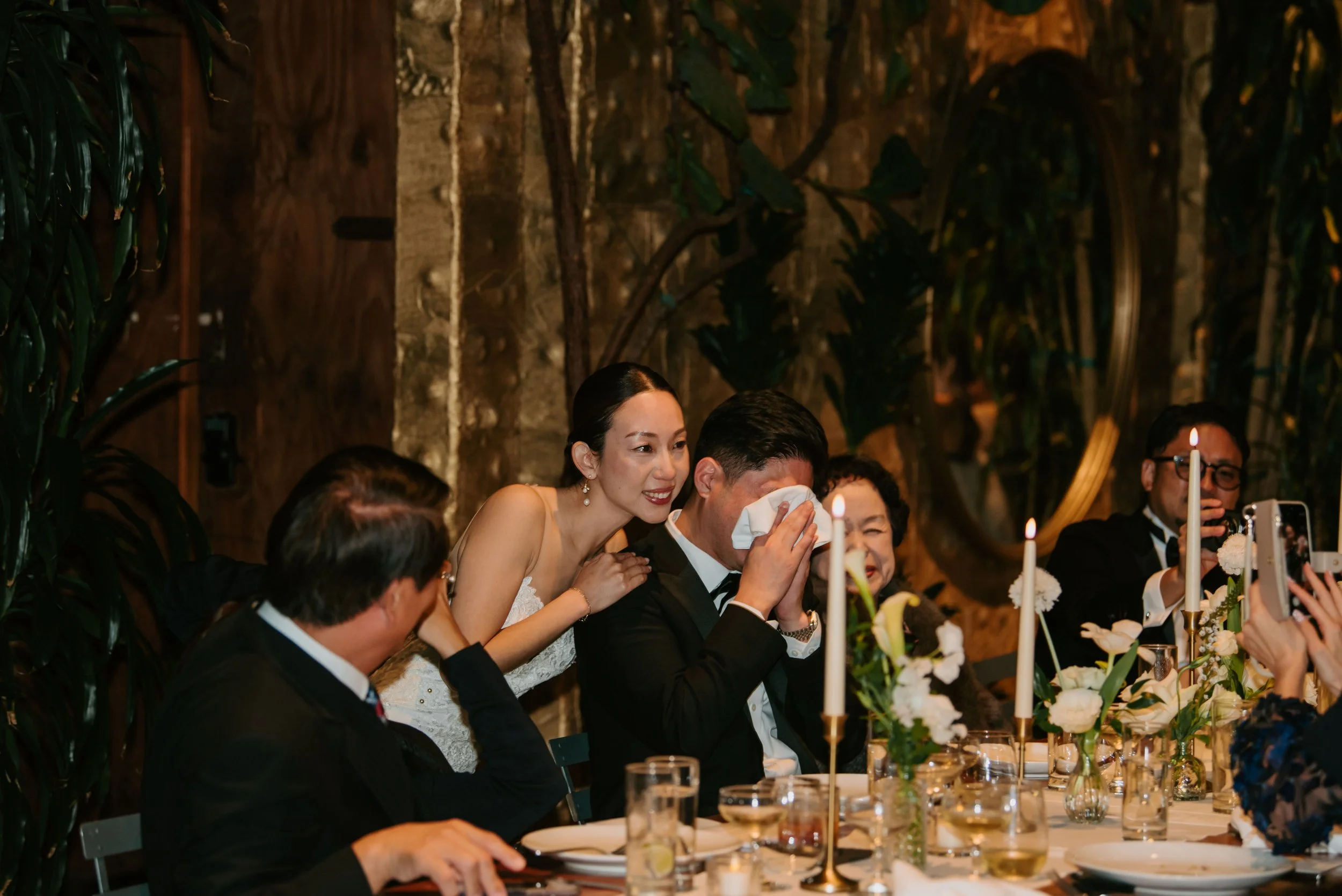 Bride's brother crying at his seat at the reception holding a napkin up to his face as the bride hugs him around the shoulders