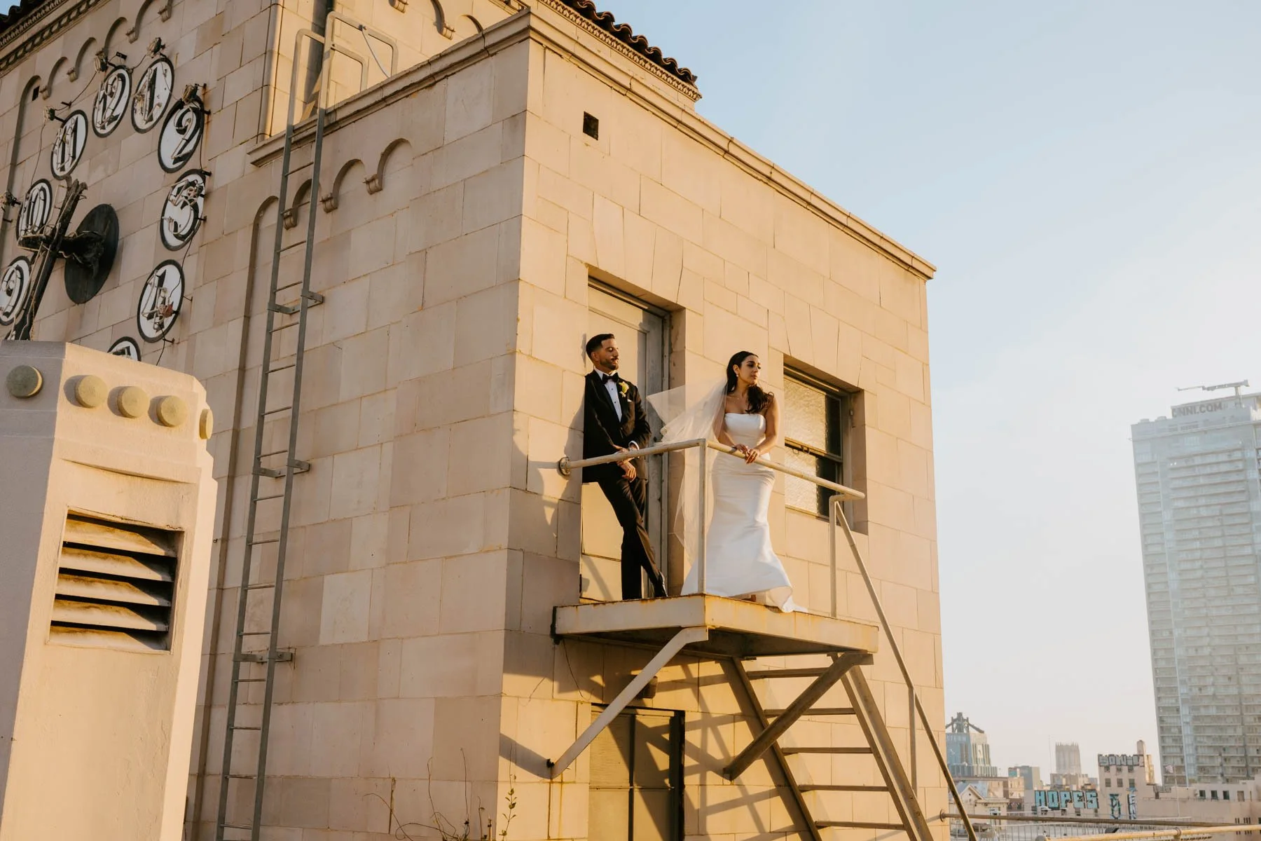 Bride and Groom standing on a little balcony. Bride is leaning against a railing look off. Groom is leaning again a door looking at bride